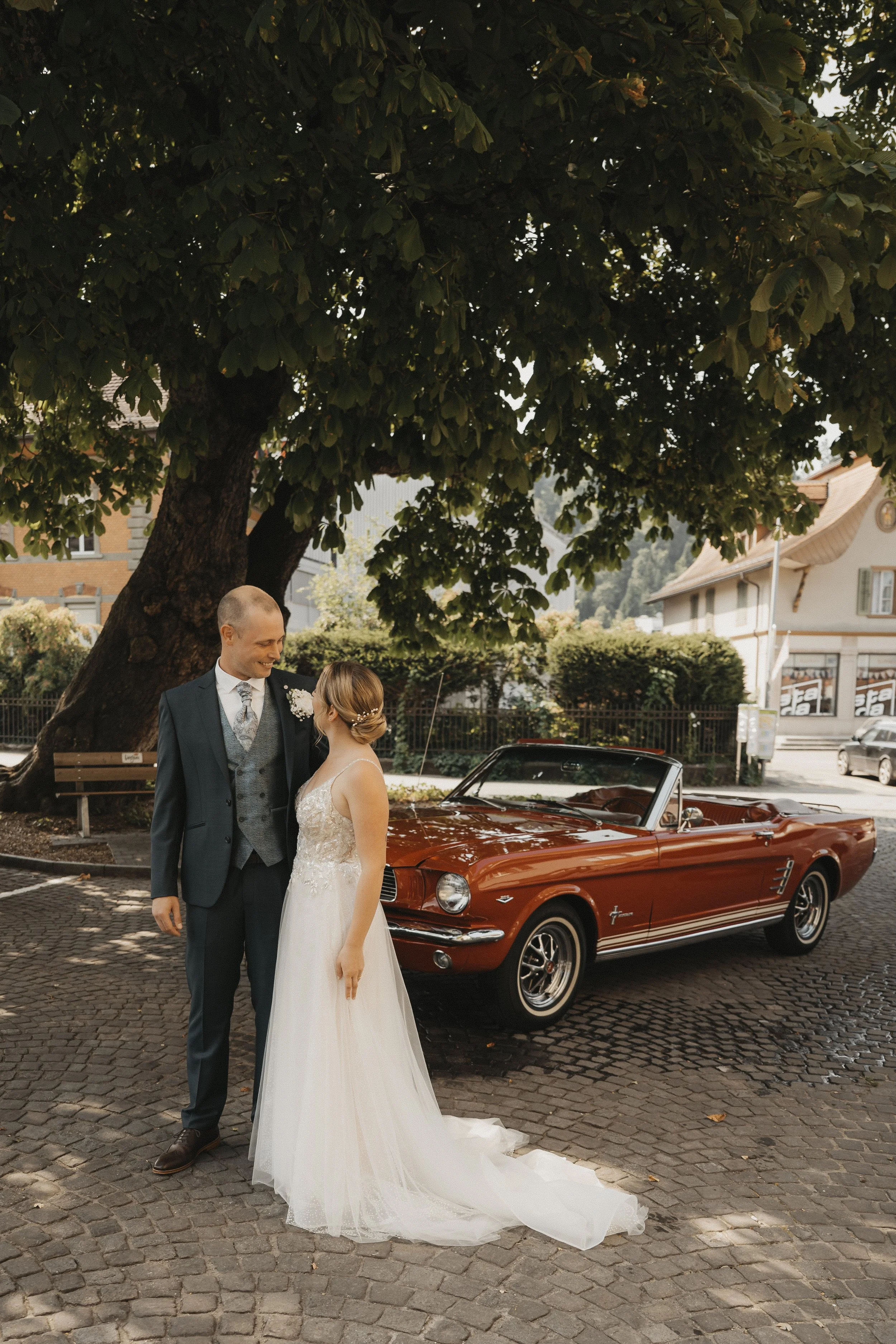 Paar in Hochzeitskleidung vor einem roten Oldtimer-Cabrio unter einem großen Baum in einer kleinen Stadt.