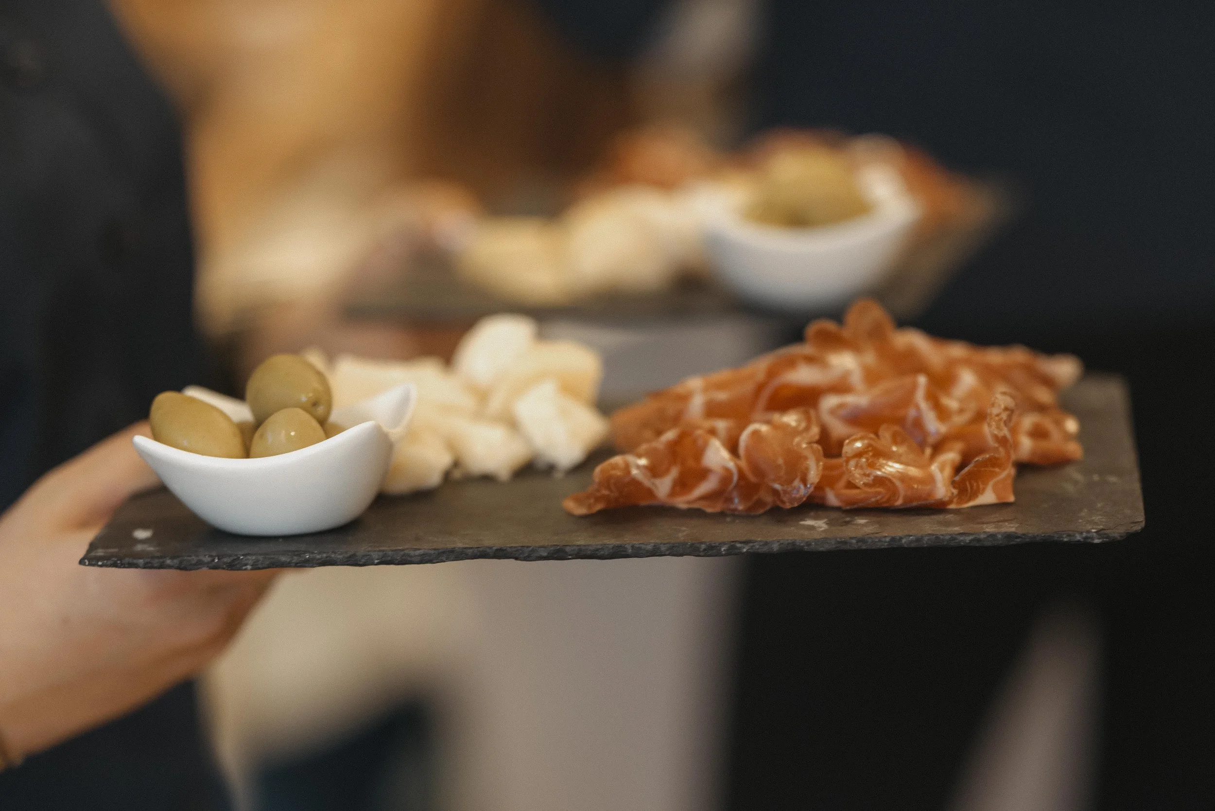 Auf einer dunklen Servierplatte liegen grüne Oliven, kleine Stücke weißer Käse und Scheiben rotem Lachs, mit zwei kleinen Schälchen mit weiteren Beilagen im Hintergrund.