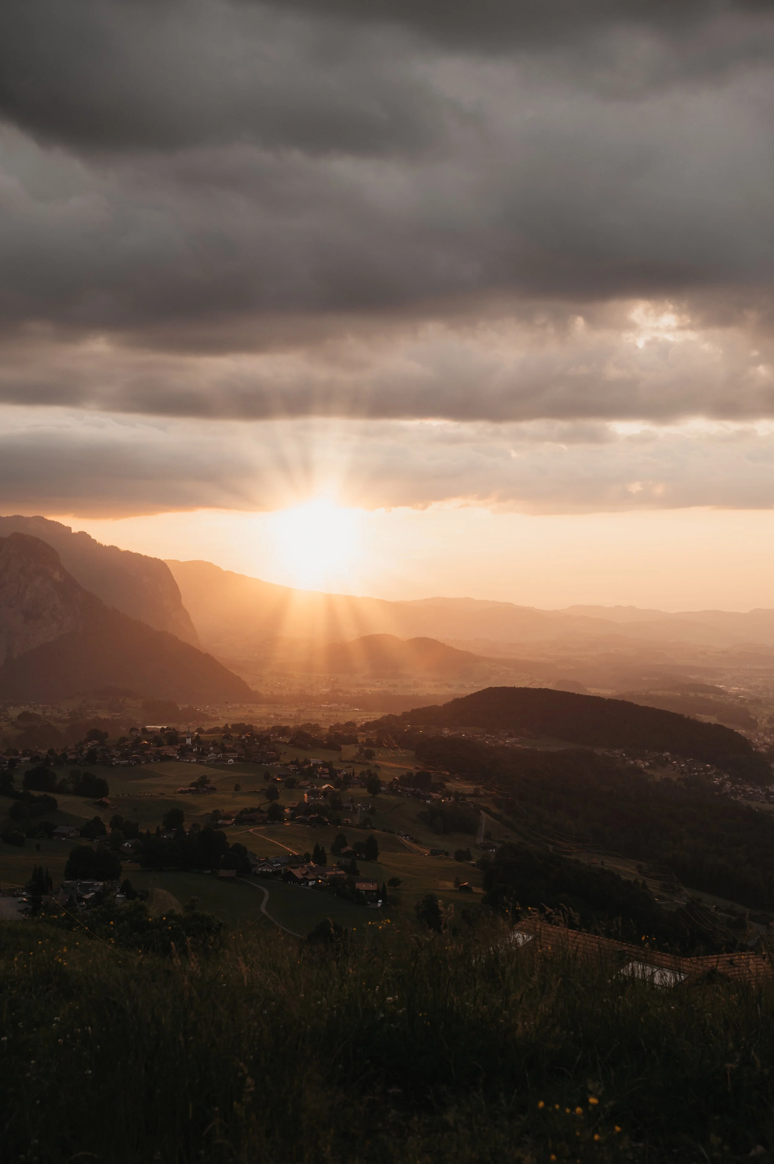 Blick auf eine ländliche Landschaft bei Sonnenuntergang mit Bergen im Hintergrund und dunklen Wolken am Himmel.