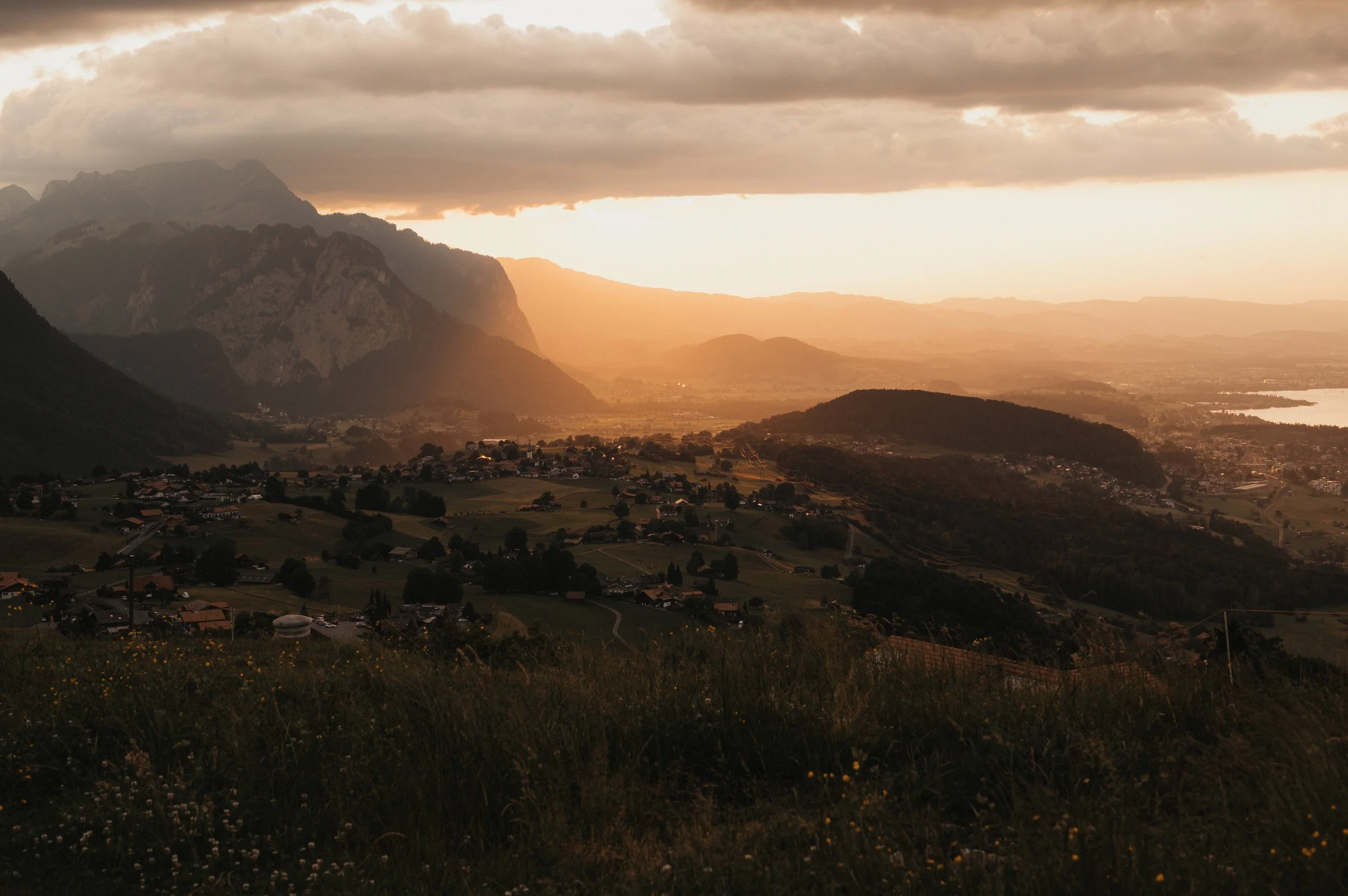 Blick auf ein Tal mit Häusern, umgeben von Bergen und Hügeln, bei Sonnenuntergang mit bewölktem Himmel.