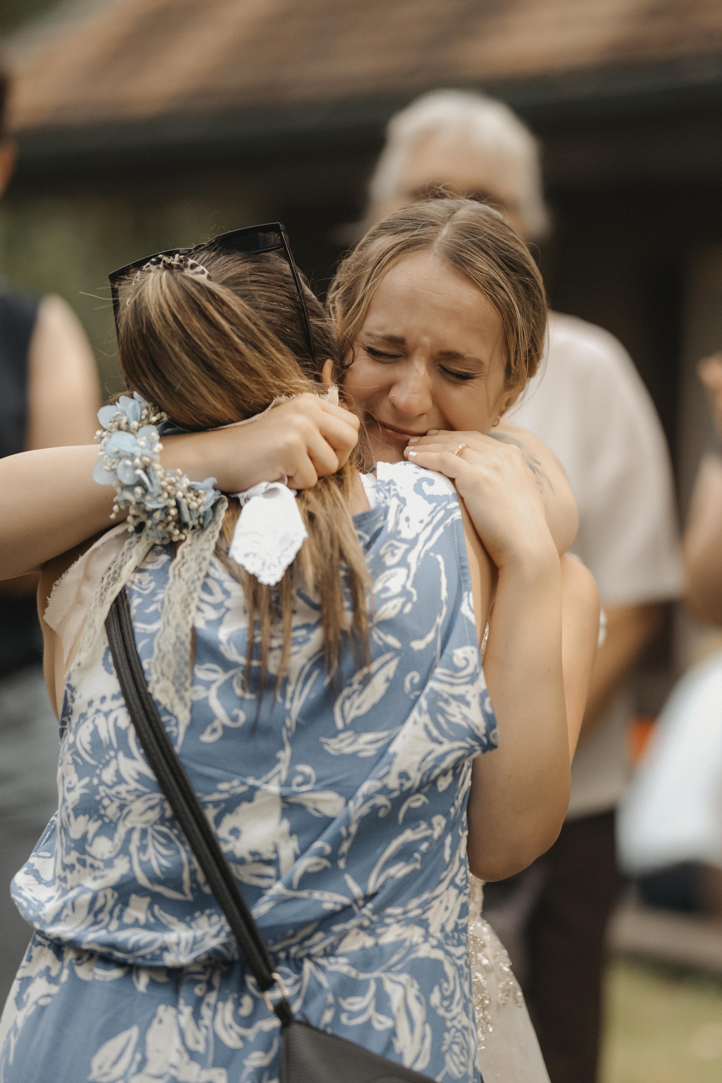 Zwei Frauen umarmen sich am Hochzeitstag, eine tristet das Brautkleid, die andere trägt ein blumiges Kleid mit Schnurrbart, alle sind emotional berührt.