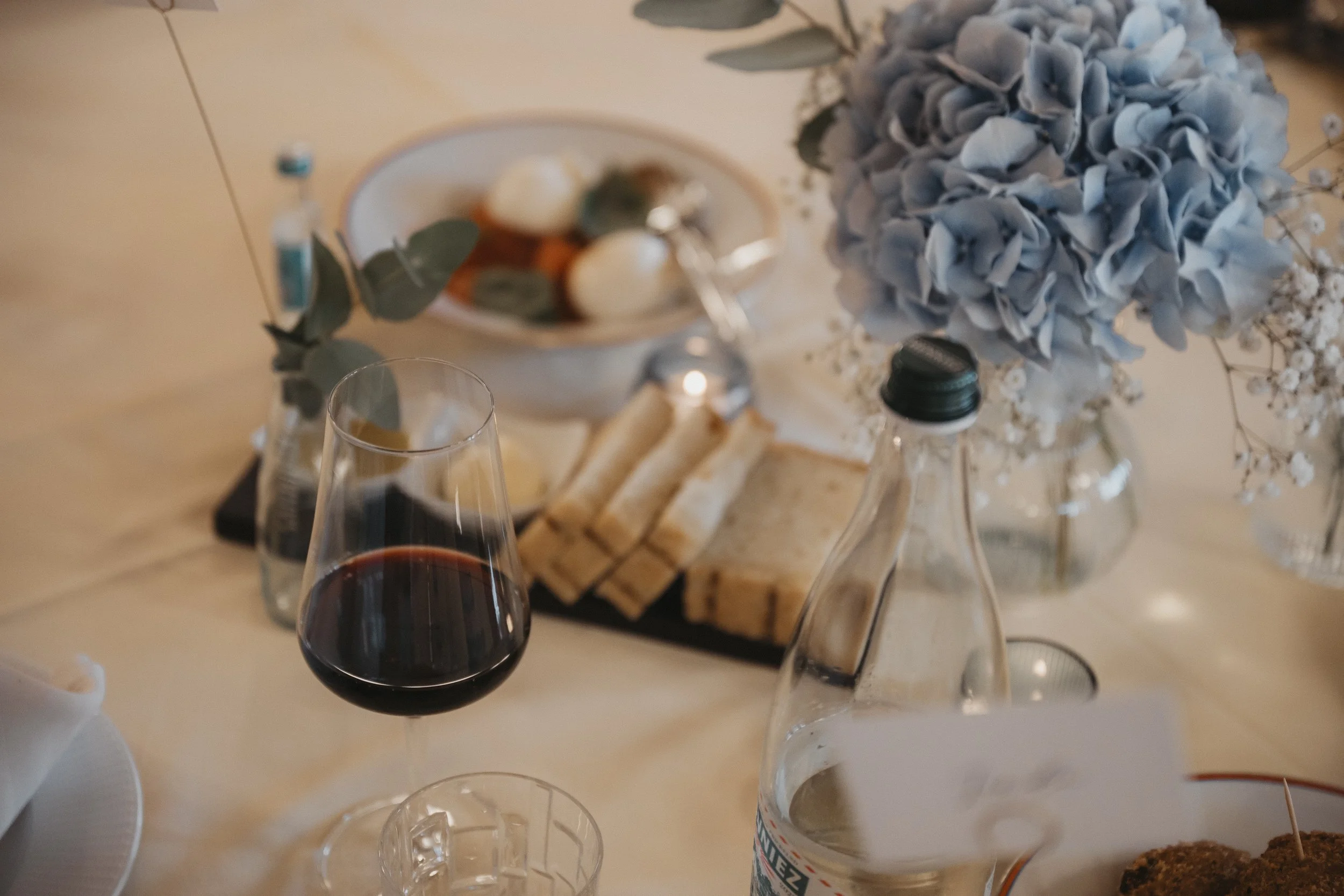 Ein gedeckter Tisch mit einem Glas Rotwein, einer Wasserflasche, einem Blumenbouquet aus blauen Hortensien, einem Teller mit Eiern, einem Butterbrot, und weiteren Flaschen. Es ist eine festliche oder elegante Mahlzeit.