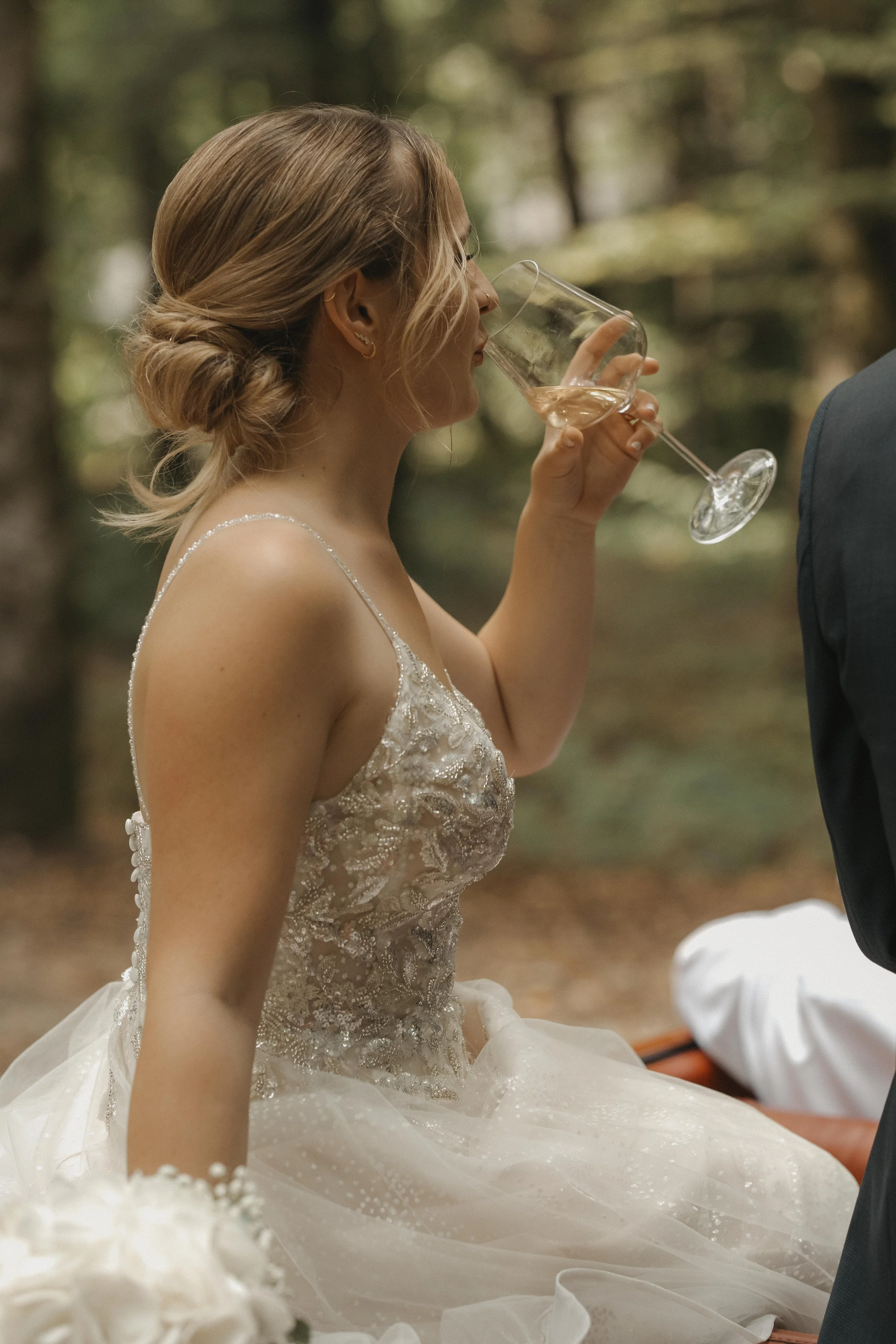 Braut in einem weißen Kleid beim Trinken aus einem Weinglas bei einer Hochzeit im Wald.