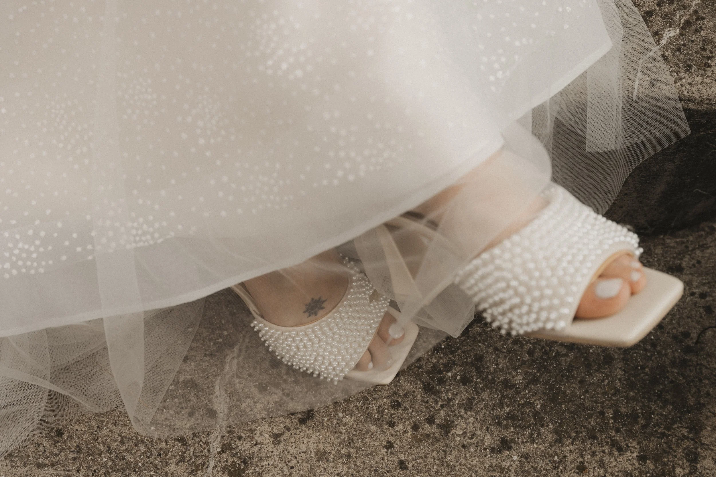 Hochzeitsschuhe mit Perlen, sichtbar unter einem weißen Brautkleid, auf einem Steinboden.