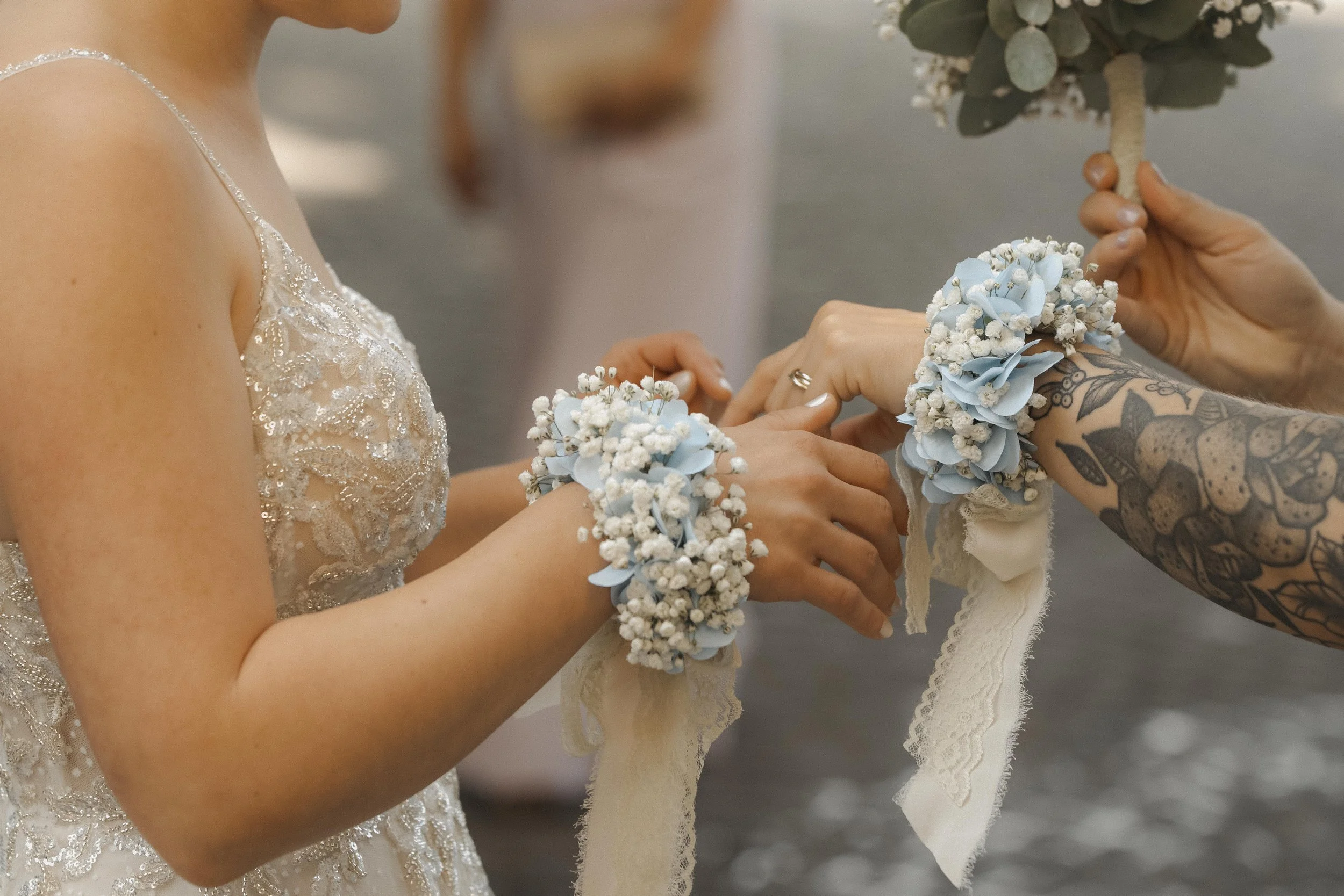 Nahaufnahme einer Hochzeitszeremonie, bei der eine Braut und eine andere Person Blumenkränze am Handgelenk anlegen, mit weiteren Blumen an einem Hochzeitsschleier. Die Person auf der rechten Seite hat Tattoos am Arm.