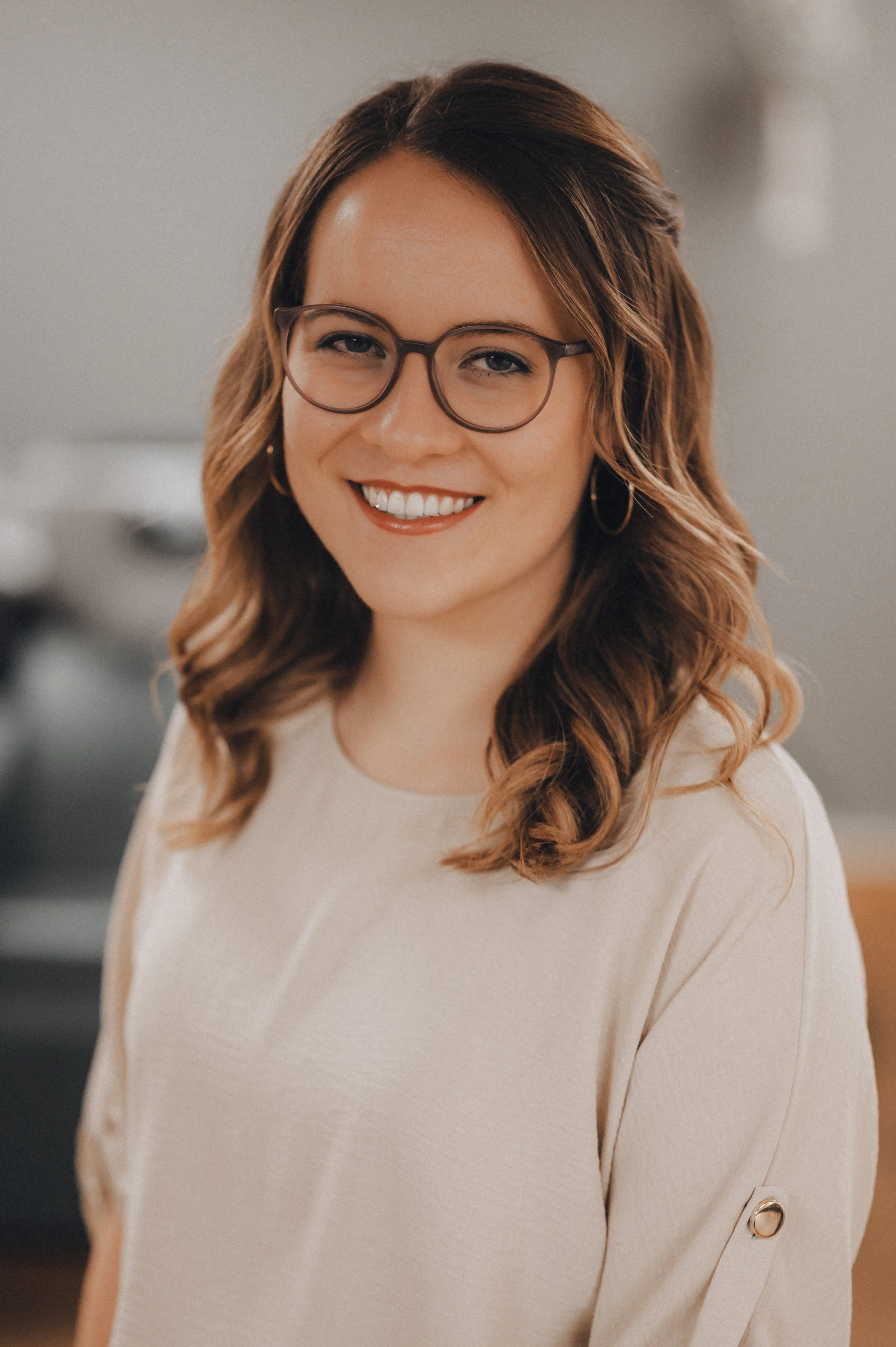 Lächelnde Frau mit Brille und Locken, trägt ein cremefarbenes Oberteil, im Hintergrund unscharf