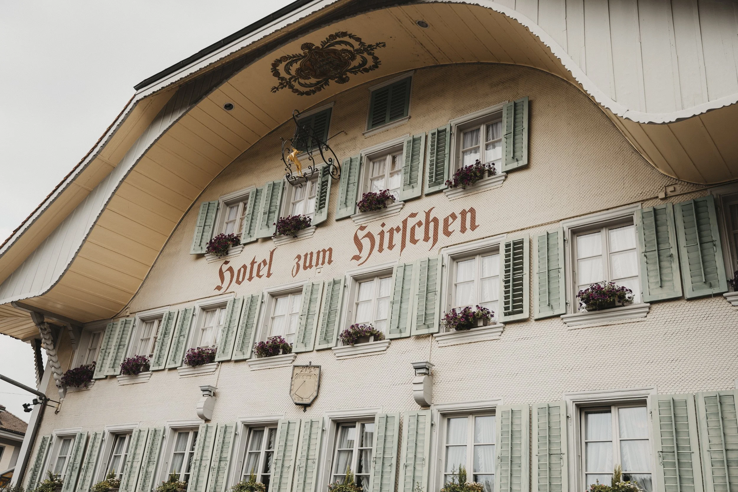 Ein mehrstöckiges Hotelgebäude mit grünen Fensterläden, Blumenkästen mit Blumen, ein Wandbild mit dem Schriftzug "Hotel zum Hirchen" und einem Eckschild.
