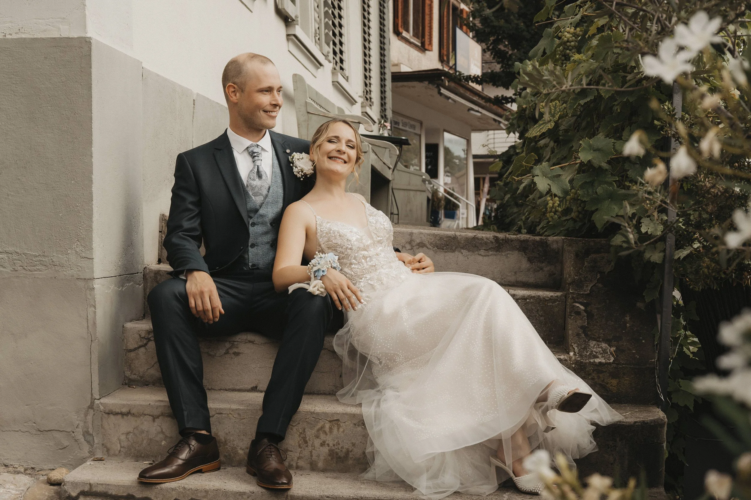 Ein frisch verheiratetes Paar sitzt lachend auf einer Treppe vor einem Haus, umgeben von Blumen.