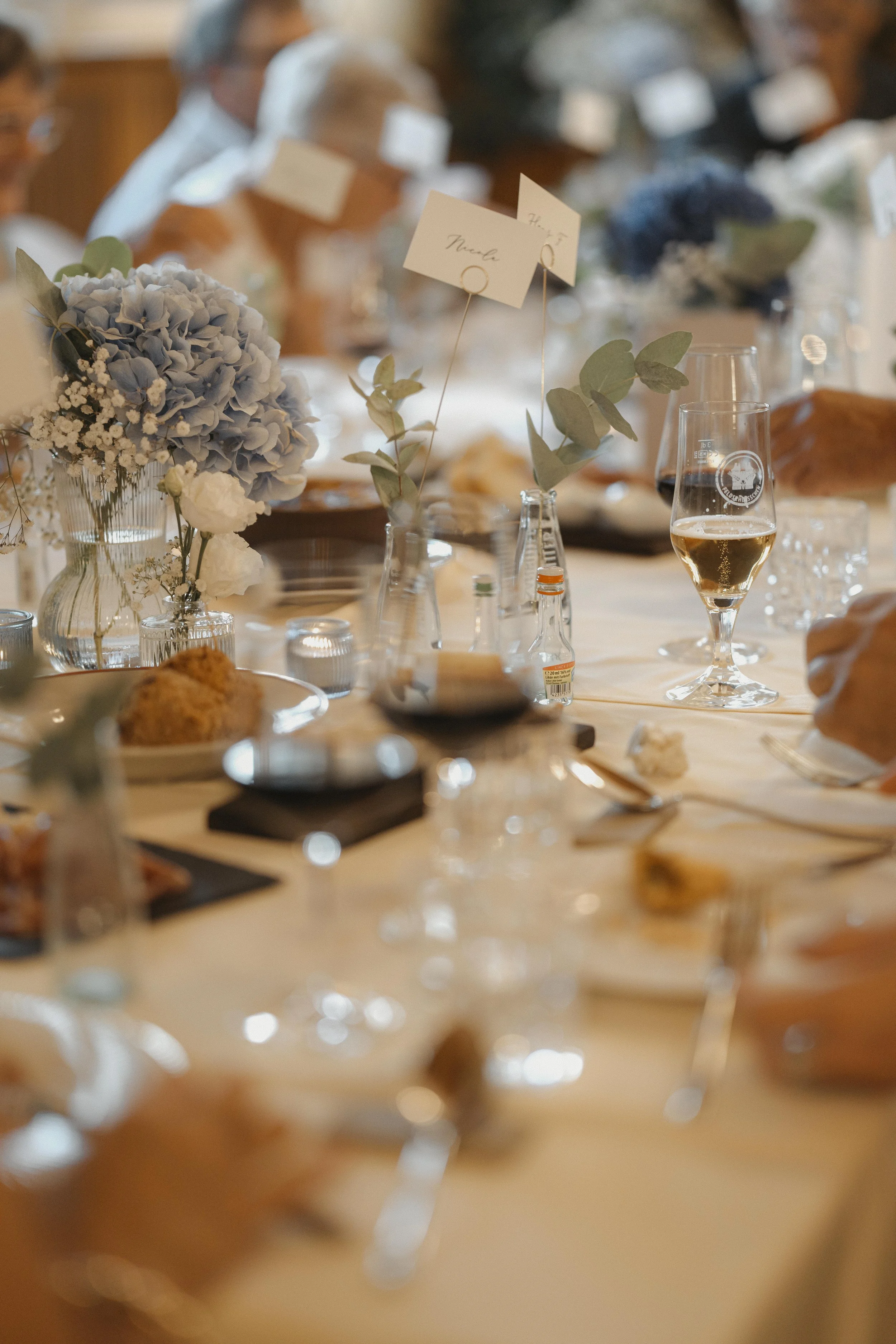 Tischdekoration bei einer festlichen Veranstaltung mit Blumen, Kerzen, Gläsern und einem Platzschild.