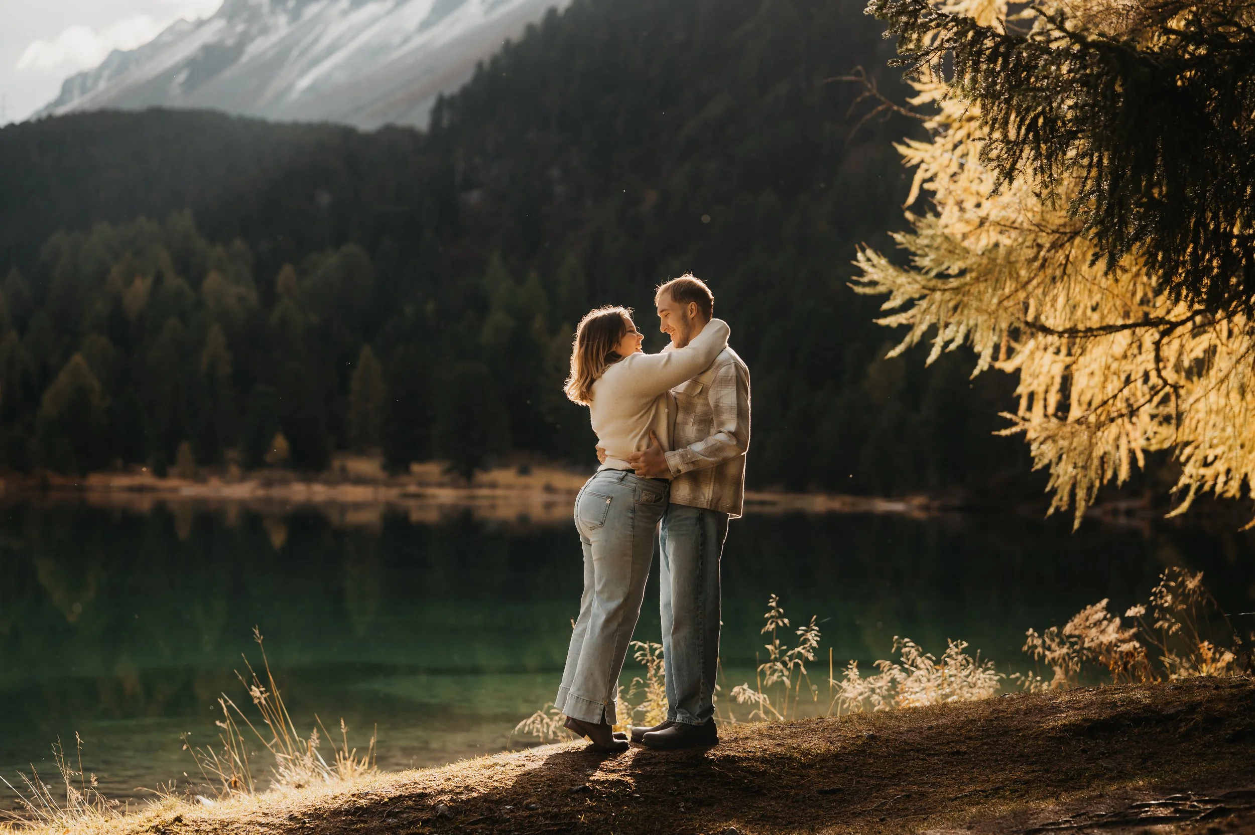 Ein Paar umarmt sich an einem See im Wald bei Sonnenuntergang, umgeben von Bäumen und Bergen.