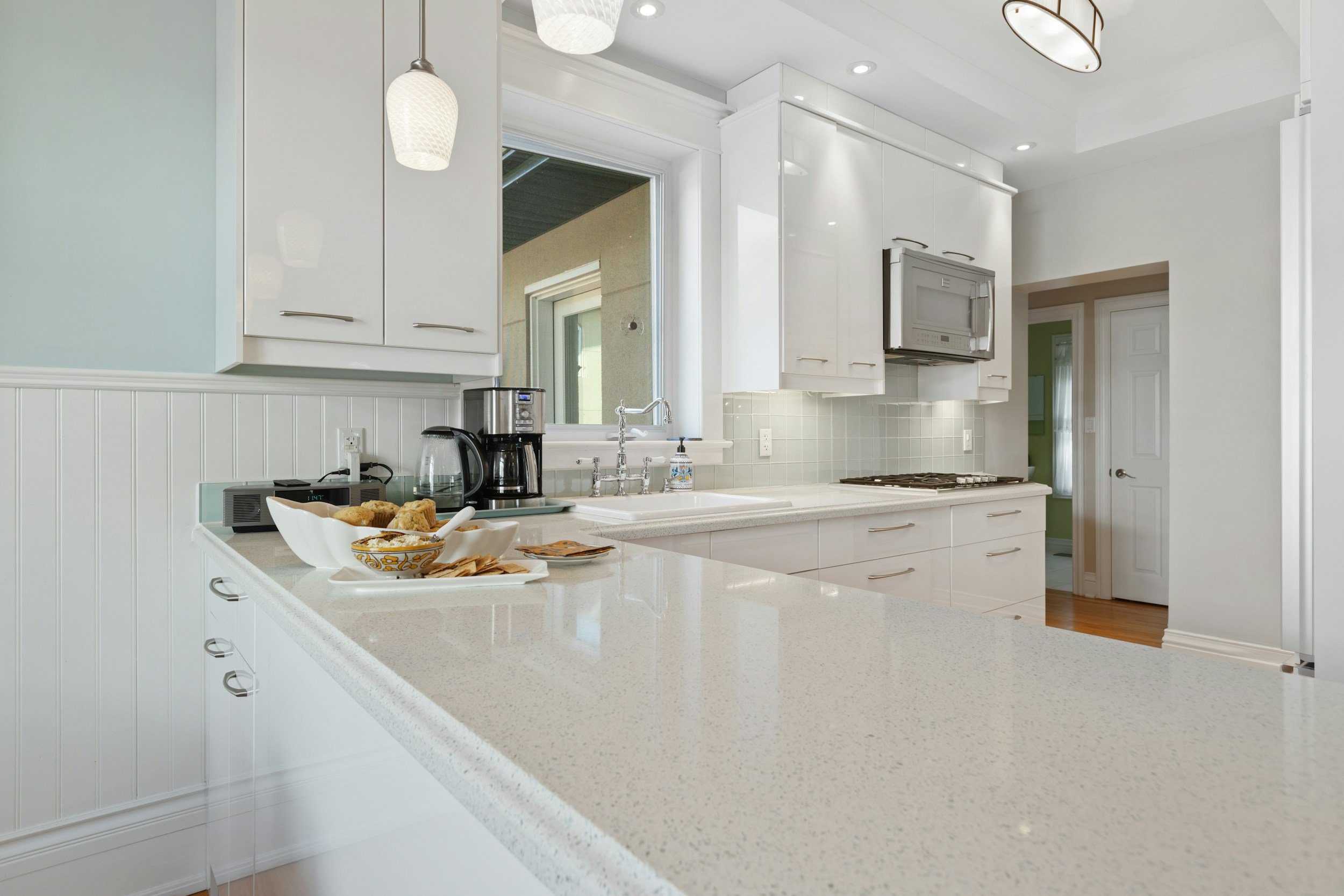 A modern kitchen with white cabinets, a white countertop, a microwave, coffee maker, and snacks on the counter.