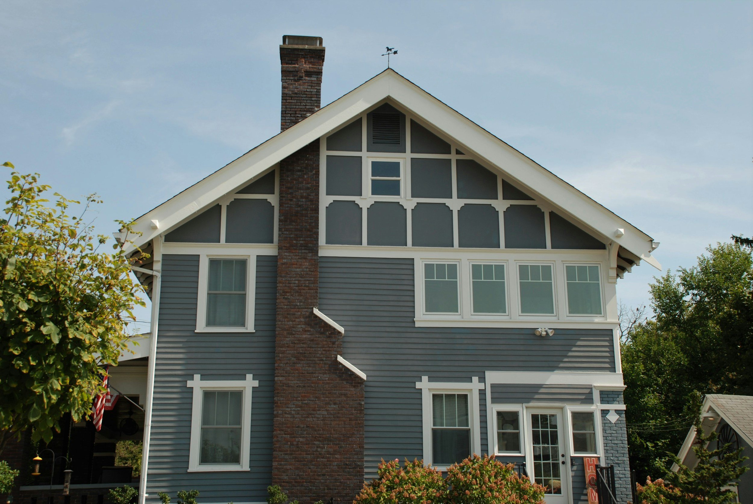 A three-story house with blue siding, white trim, and a large brick chimney. The house has multiple windows and a small porch with a door. There are trees and shrubs in the yard and a clear sky above.