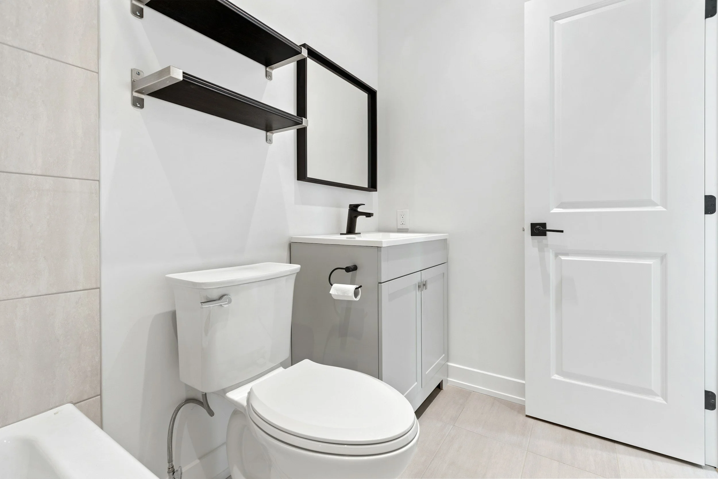 A modern bathroom with a white toilet, a grey vanity with a black faucet, a rectangular mirror, a black wall-mounted shelf, and a white door, with beige floor tiles and white painted walls.