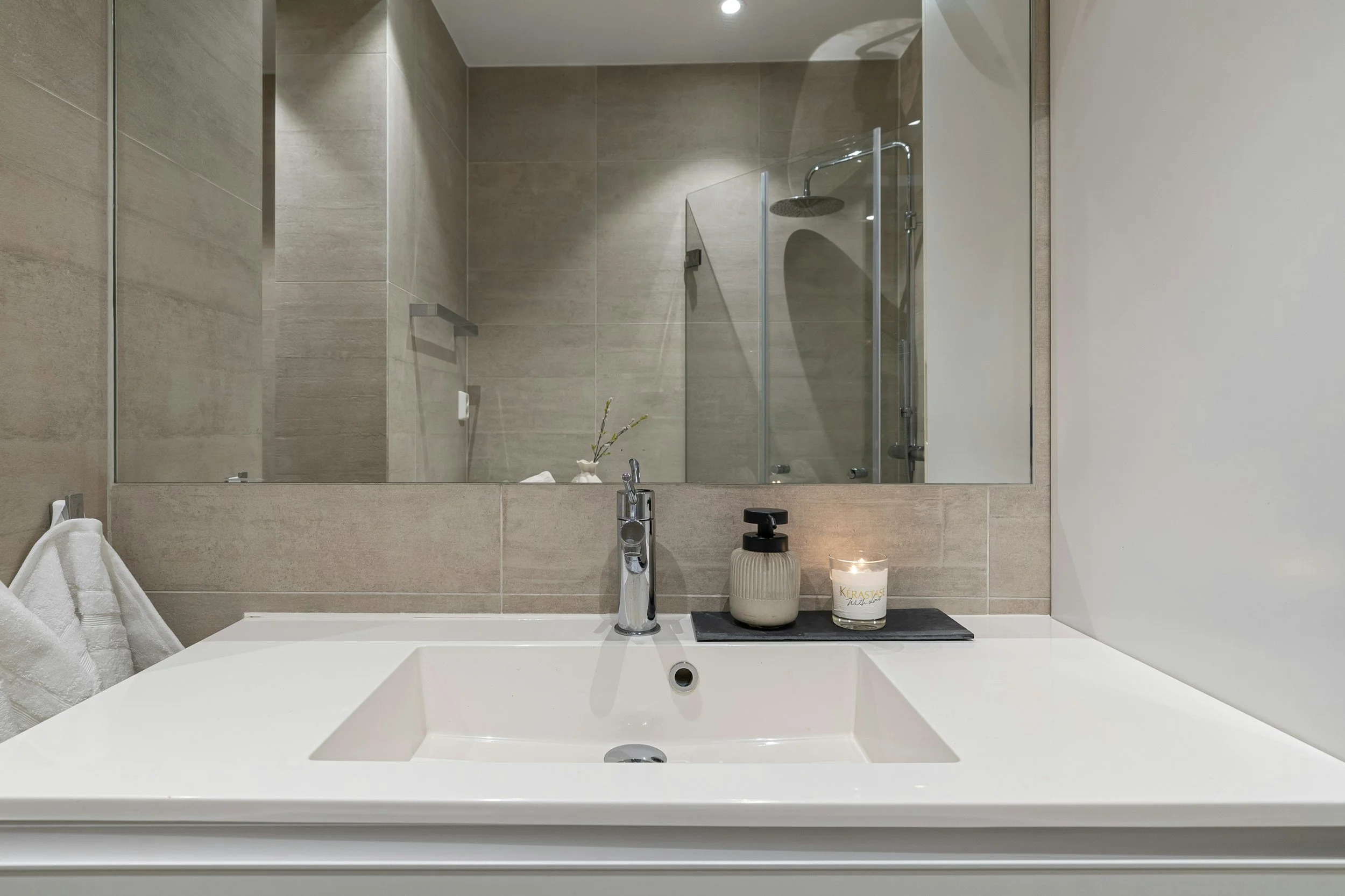 Bathroom vanity with a white sink, a modern chrome faucet, a candle, a soap dispenser, and a large mirror above the sink. In the reflection, a shower with a glass door and showerhead can be seen.