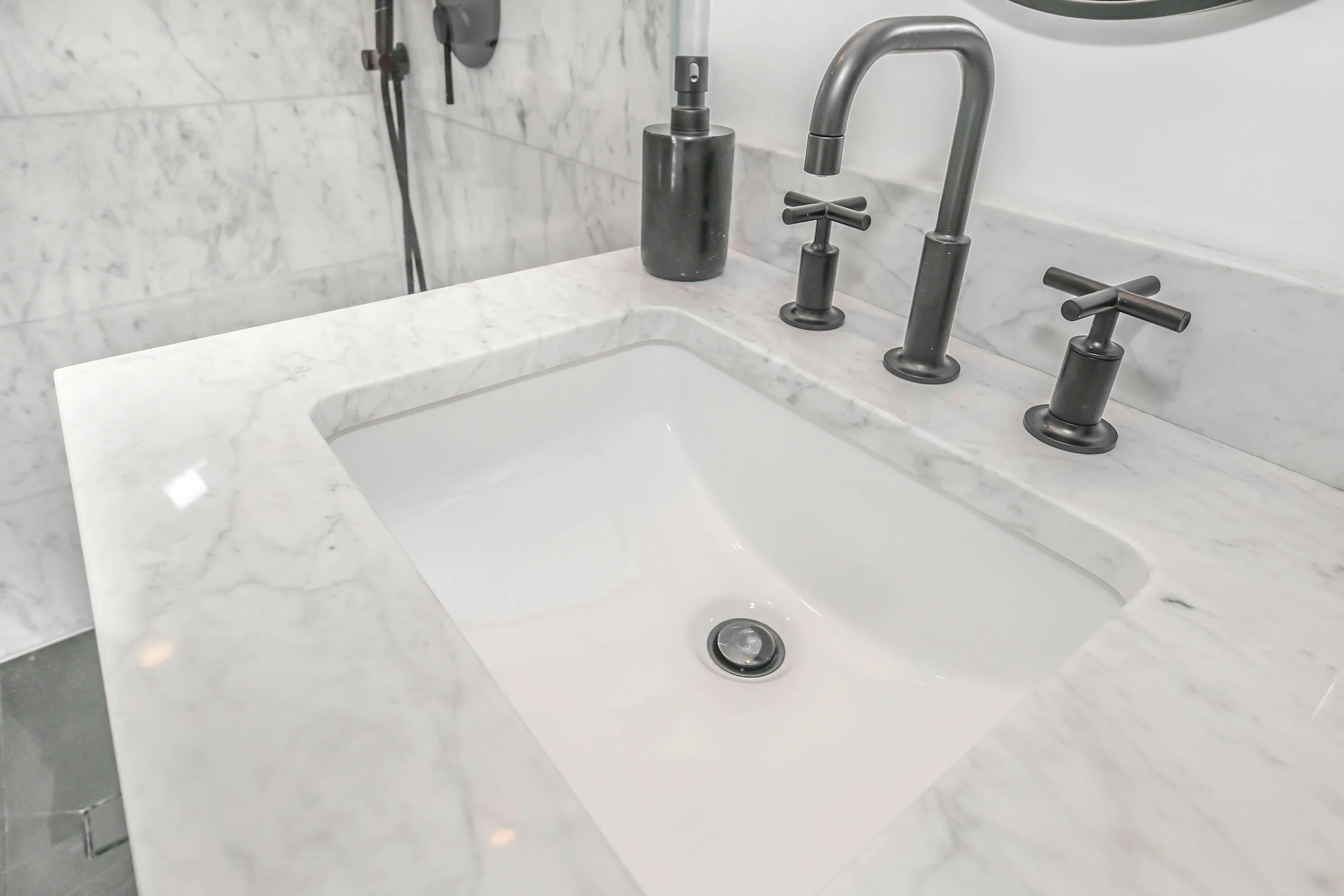 Close-up of a white marble bathroom sink with two black soap dispensers and brass fixtures, with marble walls and a mirror visible in the background.
