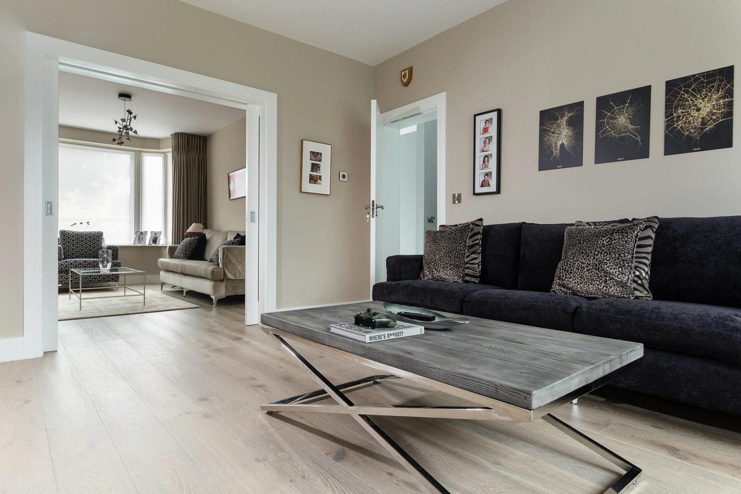 Living room with black sofa, decorative cushions, wooden coffee table with magazines, remote, and game controller, and wall art. Adjacent room with beige sofa, patterned armchair, and window seat with pillows.