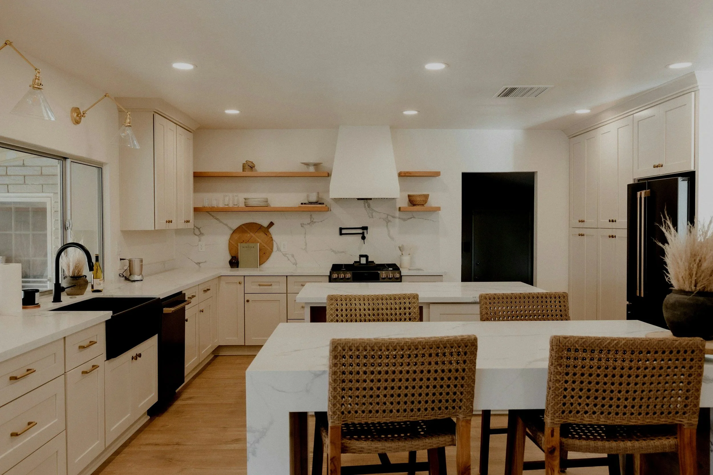 A modern kitchen with white cabinets, a white marble island, open wooden shelves, and wicker chairs around the island.