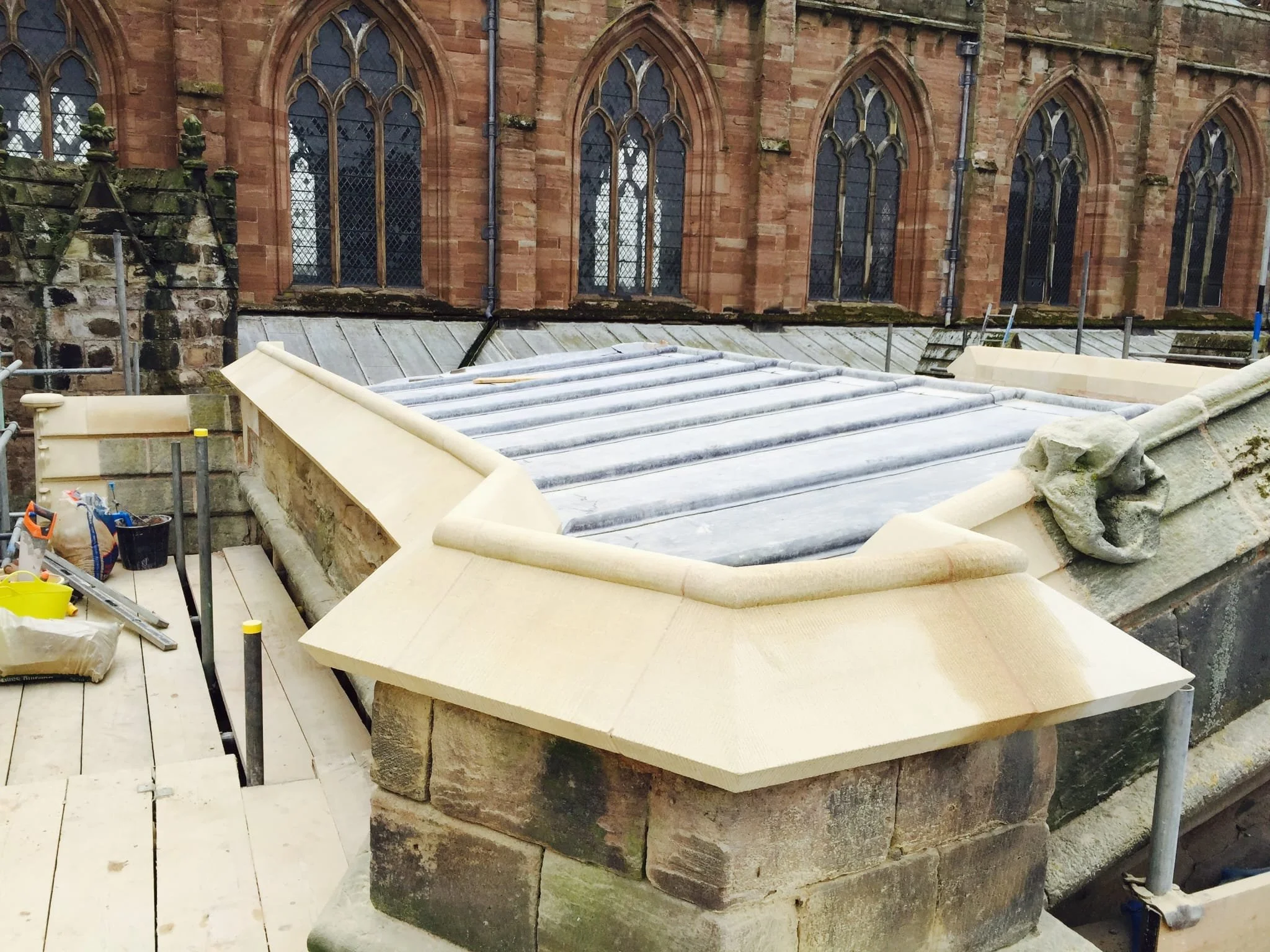 Construction site on a roof with stone and woodwork, scaffolding, tools, and a historic church with stained glass windows in the background.