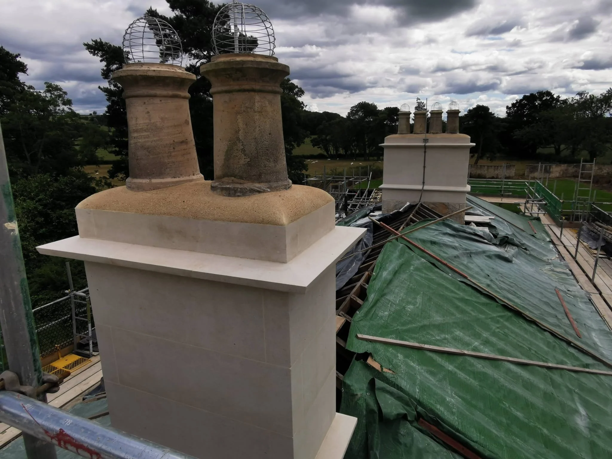 Rooftop chimney stacks under construction with scaffolding and green tarpaulin, surrounding a scenic landscape with trees and cloudy sky.
