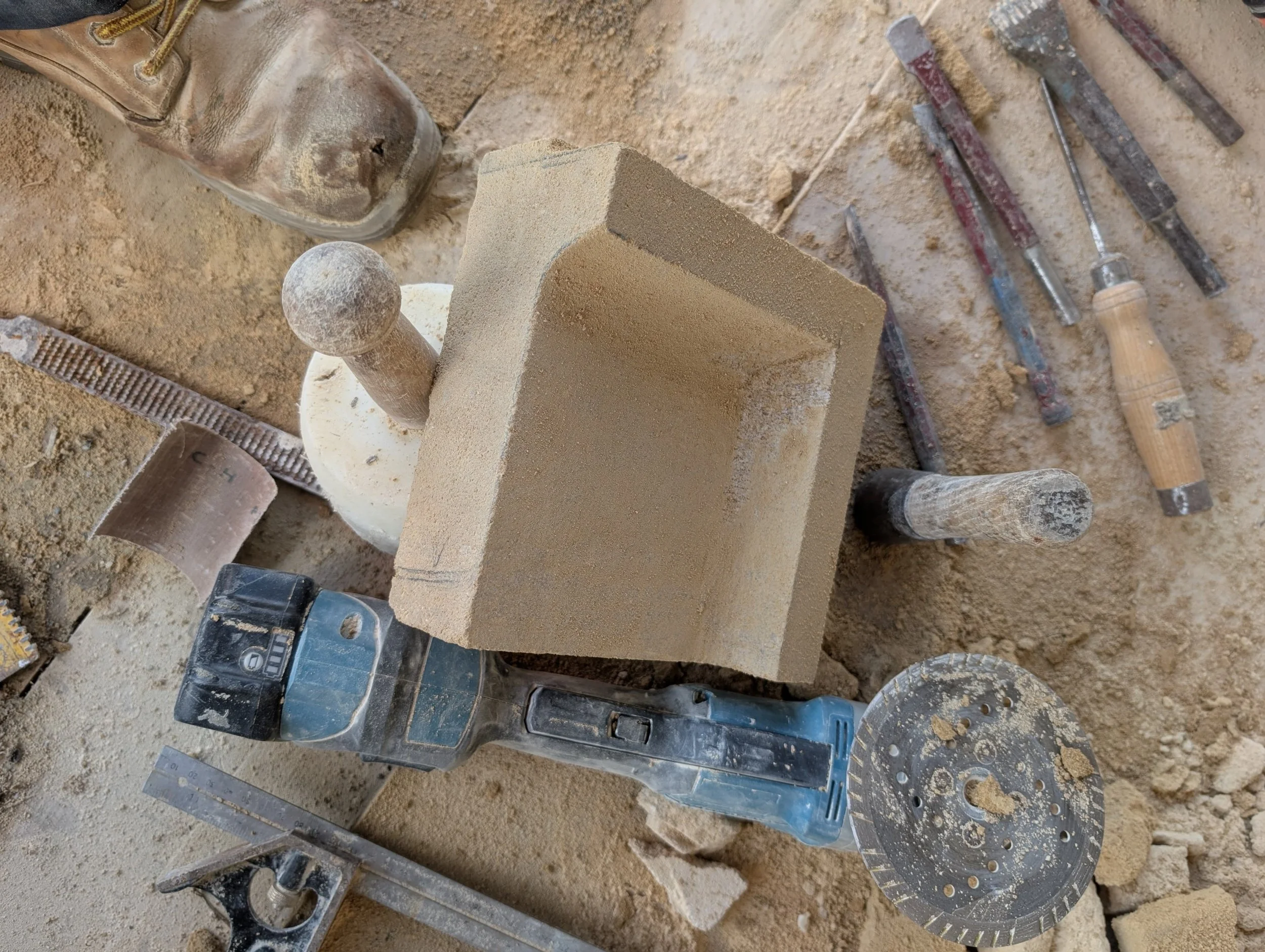 Construction site with a brick held in place by a power tool, surrounded by various chisels, scrapers, and dust on the dusty ground.