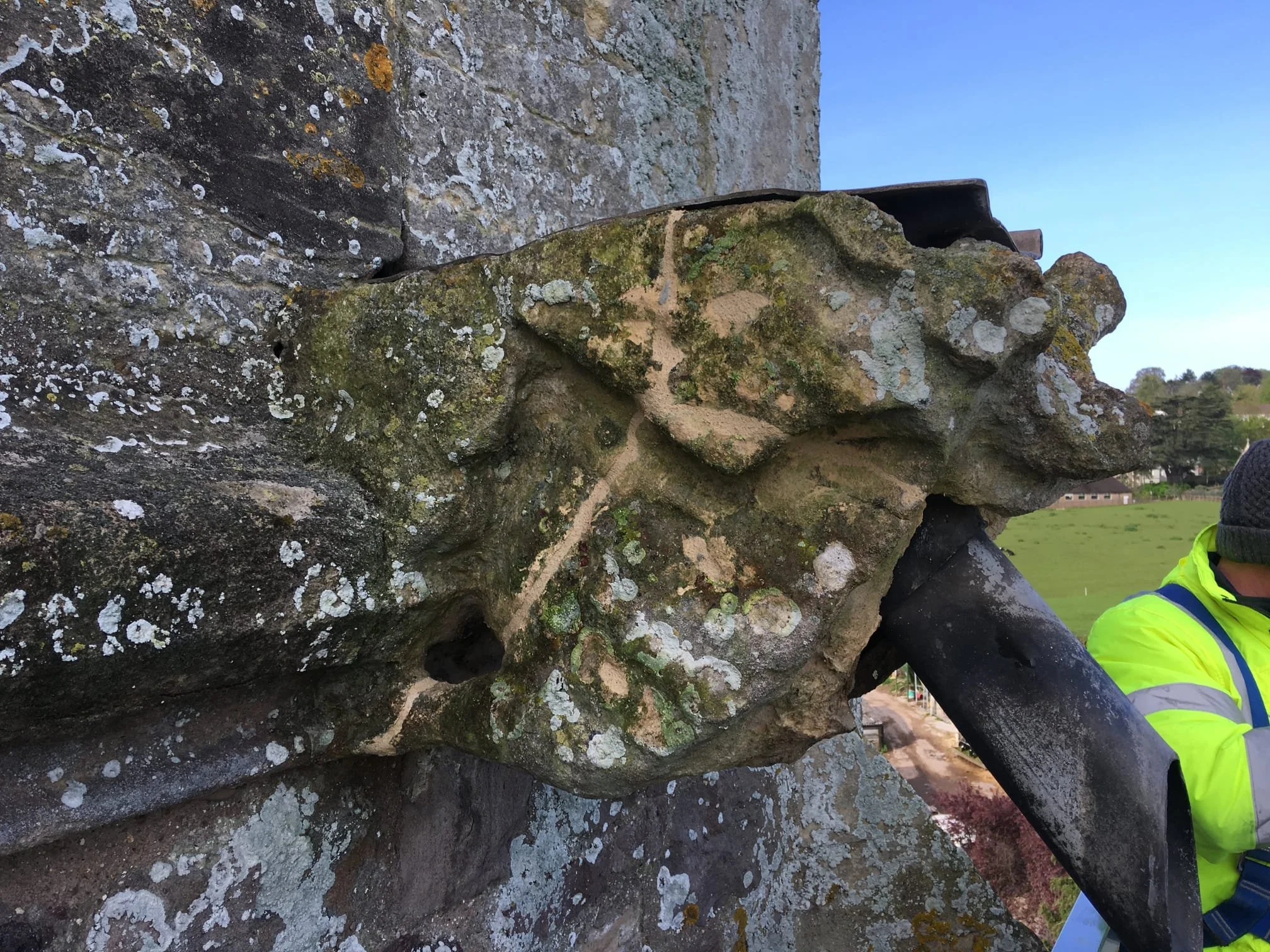 Close-up of a rock climbing hold attached to a stone climbing wall, with a person wearing a neon yellow jacket and a black cap visible in the background.