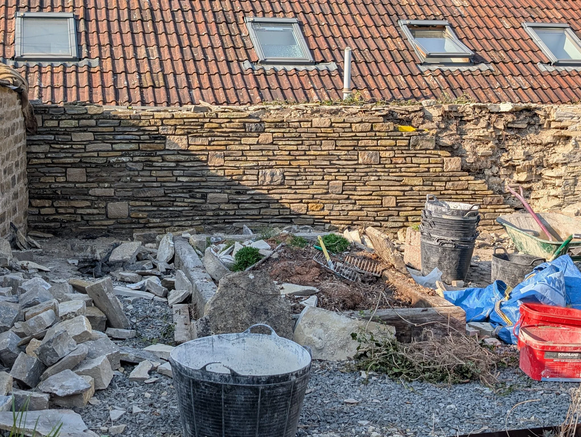 Construction site with a partially built stone wall, rubble, buckets, gardening tools, and construction materials, with a roof with skylights in the background.