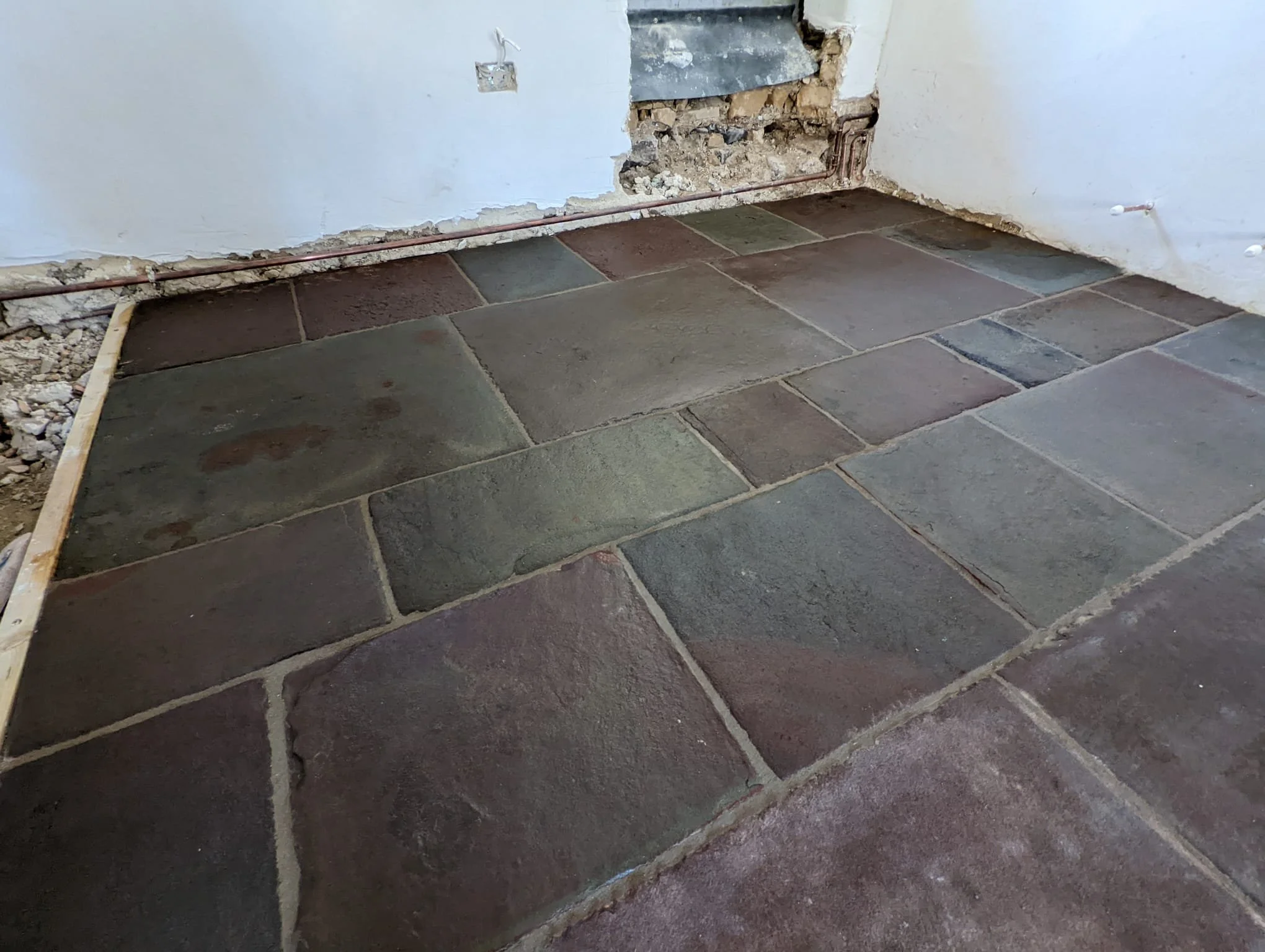 A room with partially installed slate tile flooring and an unfinished white wall with a hole. Exposed electrical wiring and plumbing pipes are visible along the wall.