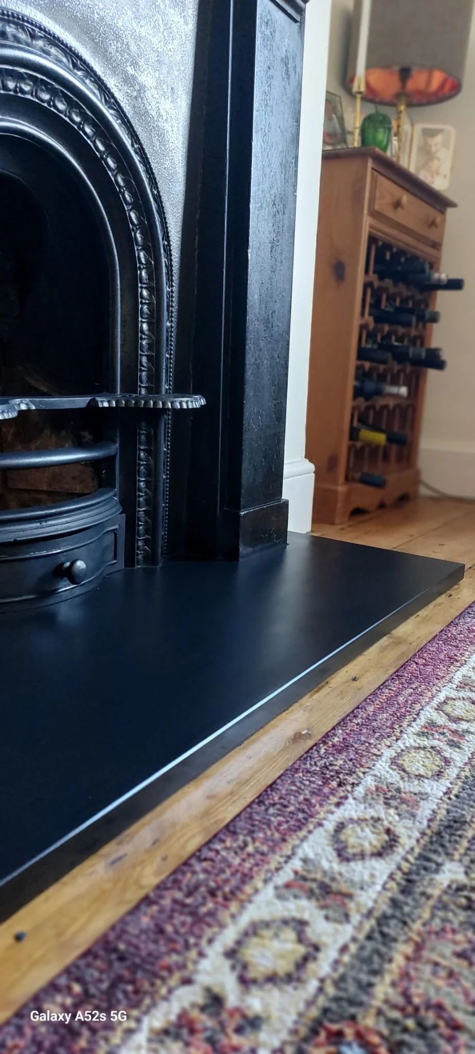 Close-up of a traditional black fireplace with a decorative metallic door and a small round drawer, positioned on a hardwood floor with a colorful patterned area rug. In the background, there's a wooden wine rack, a lamp with a red shade, and framed pictures on a mantel or shelf.