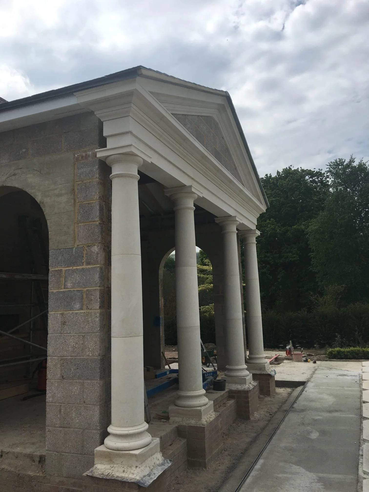 Construction site of a building with classical architecture elements, including four large, white columns with bases and capitals, supporting a triangular pediment with decorative molding, with other construction materials and equipment around.