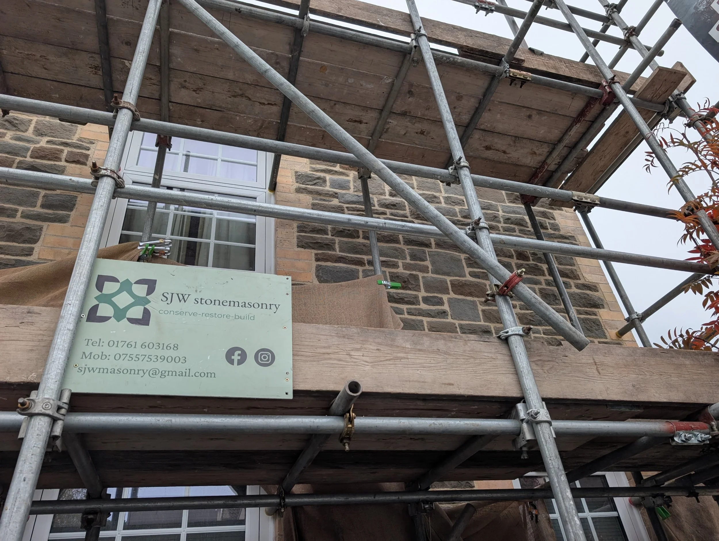 Construction scaffolding around a brick house with a sign for SJW Stonemasonry, showing their contact details and social media icons.