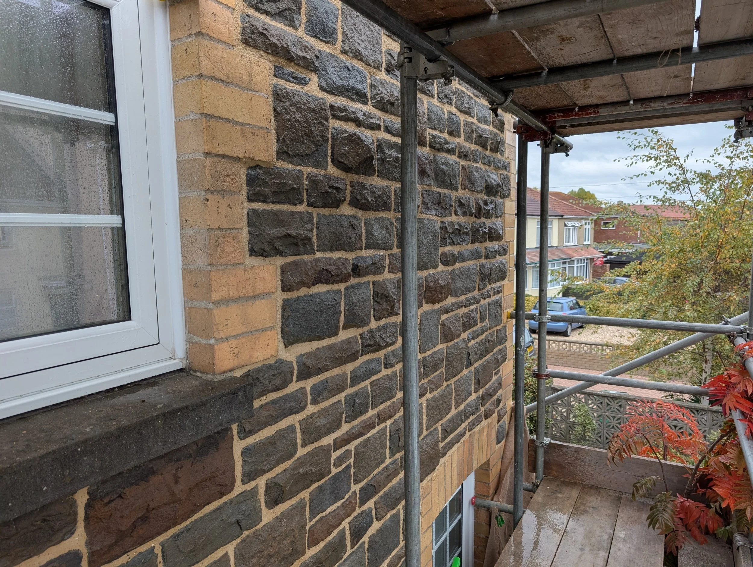 View of a residential brick house from a balcony with scaffolding, rain at window, weathered tree and neighboring houses in the background.