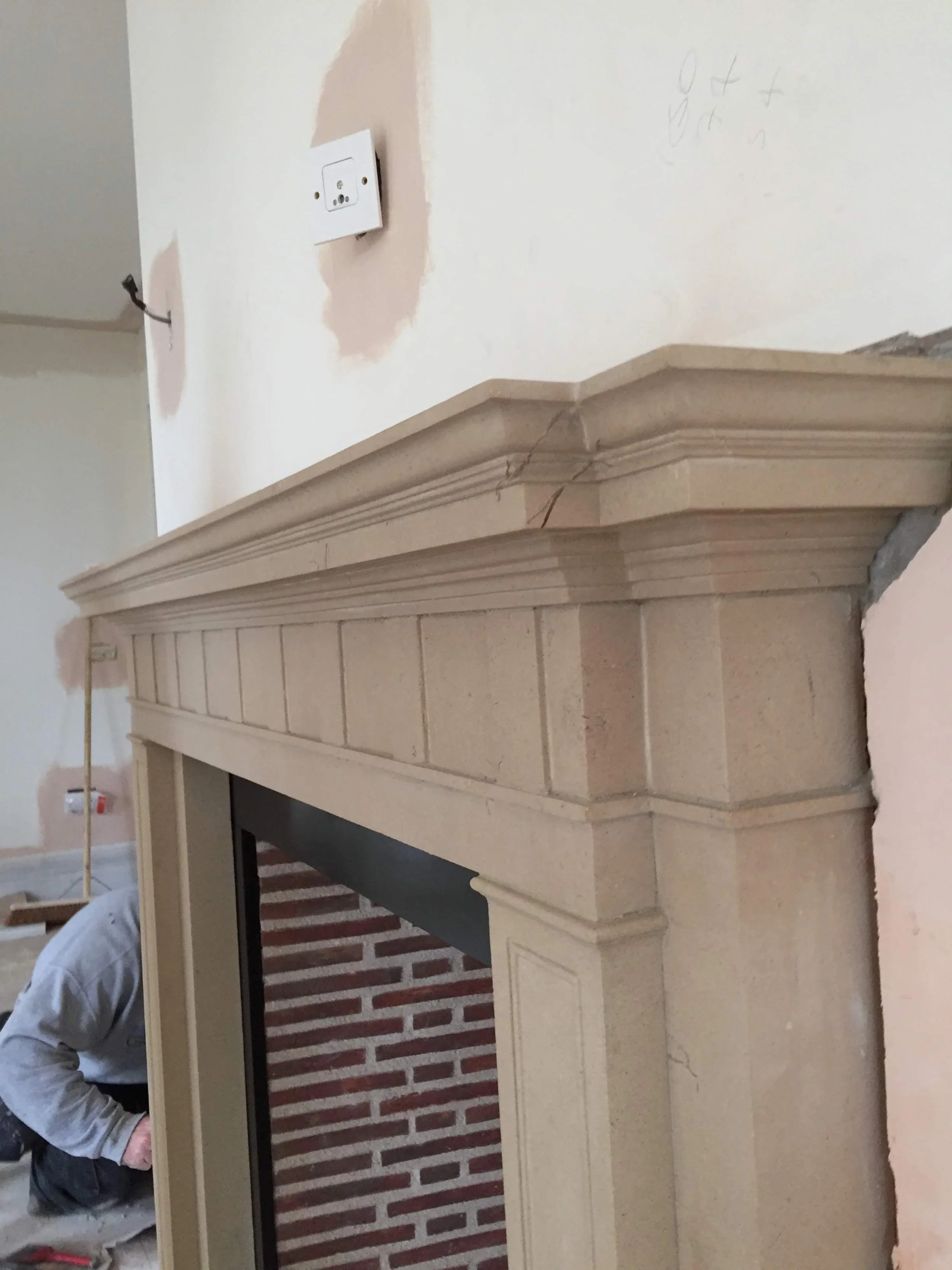 A beige fireplace mantel under construction in a room with unfinished wall and electrical outlet.