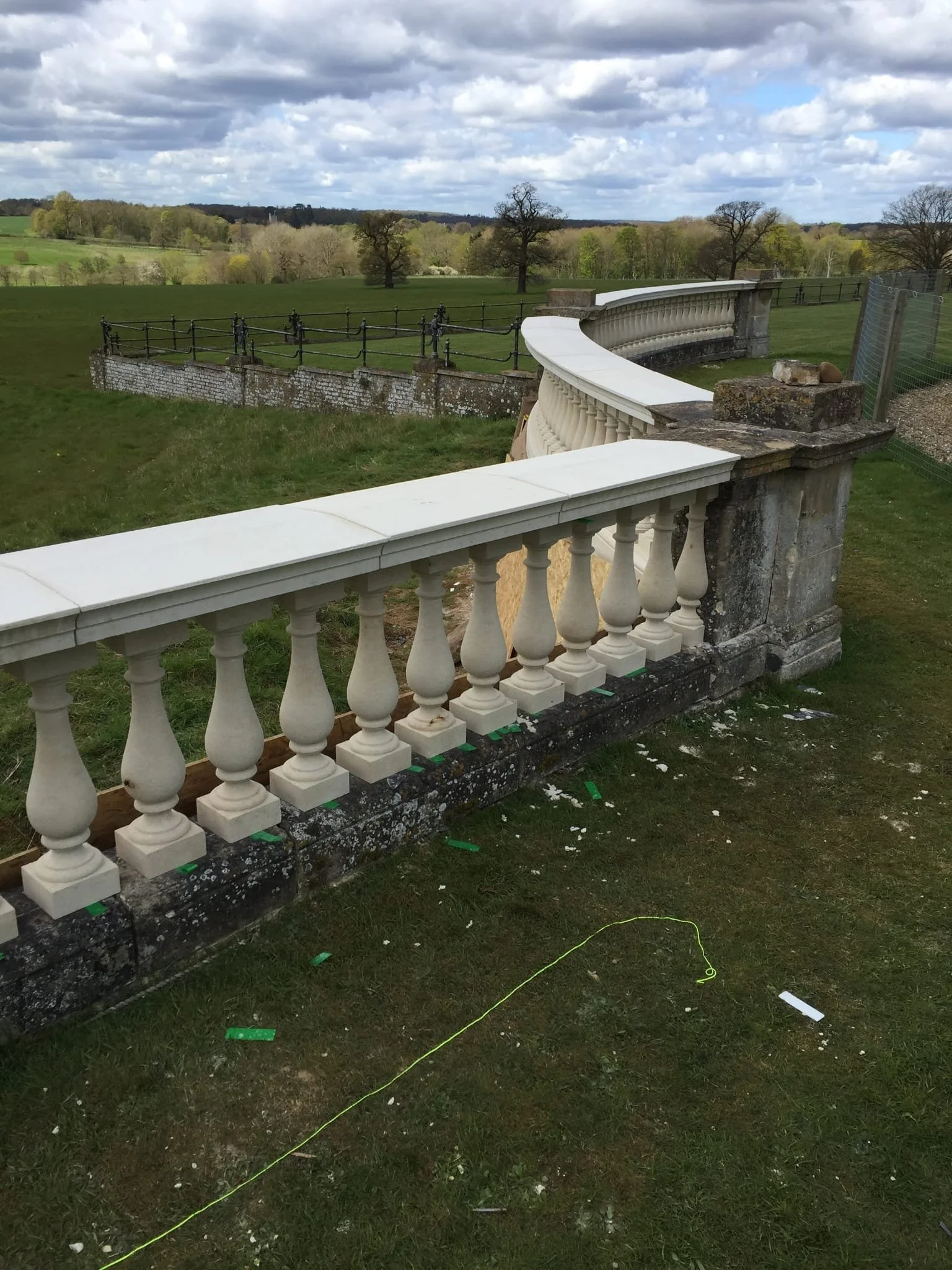 Stone and concrete balustrade railing on a grassy outdoor terrace overlooking a green field with trees in the distance, cloudy sky overhead.