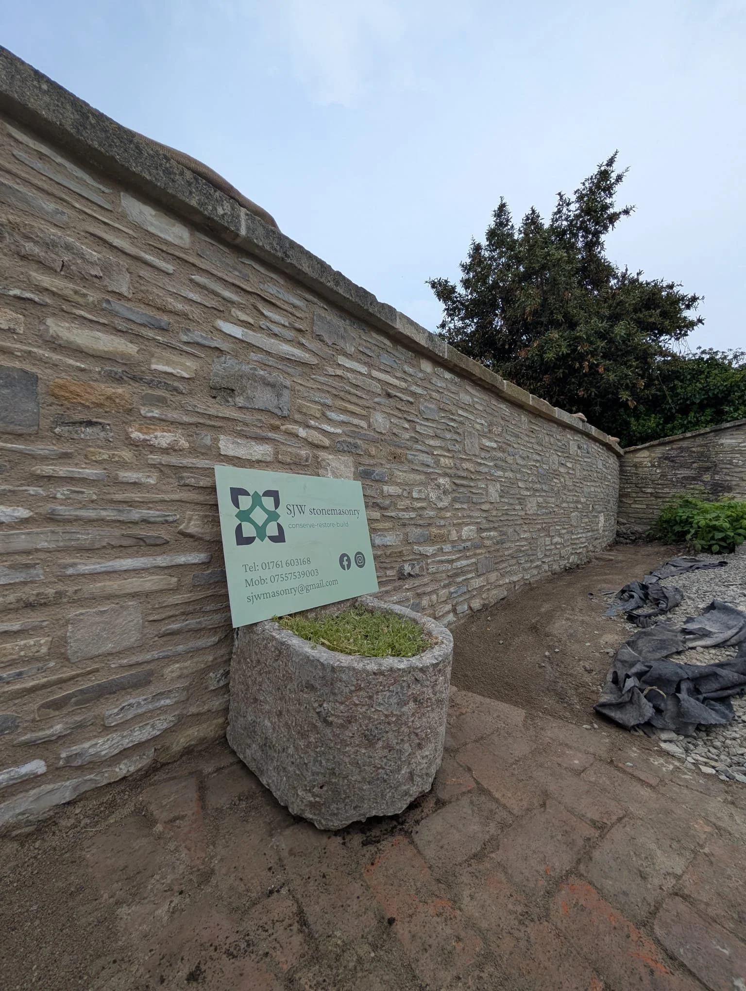 A stone wall with a business sign for SJW stonemasonry resting on a large stone planter with green grass. Construction debris and gravel are visible on the ground nearby.