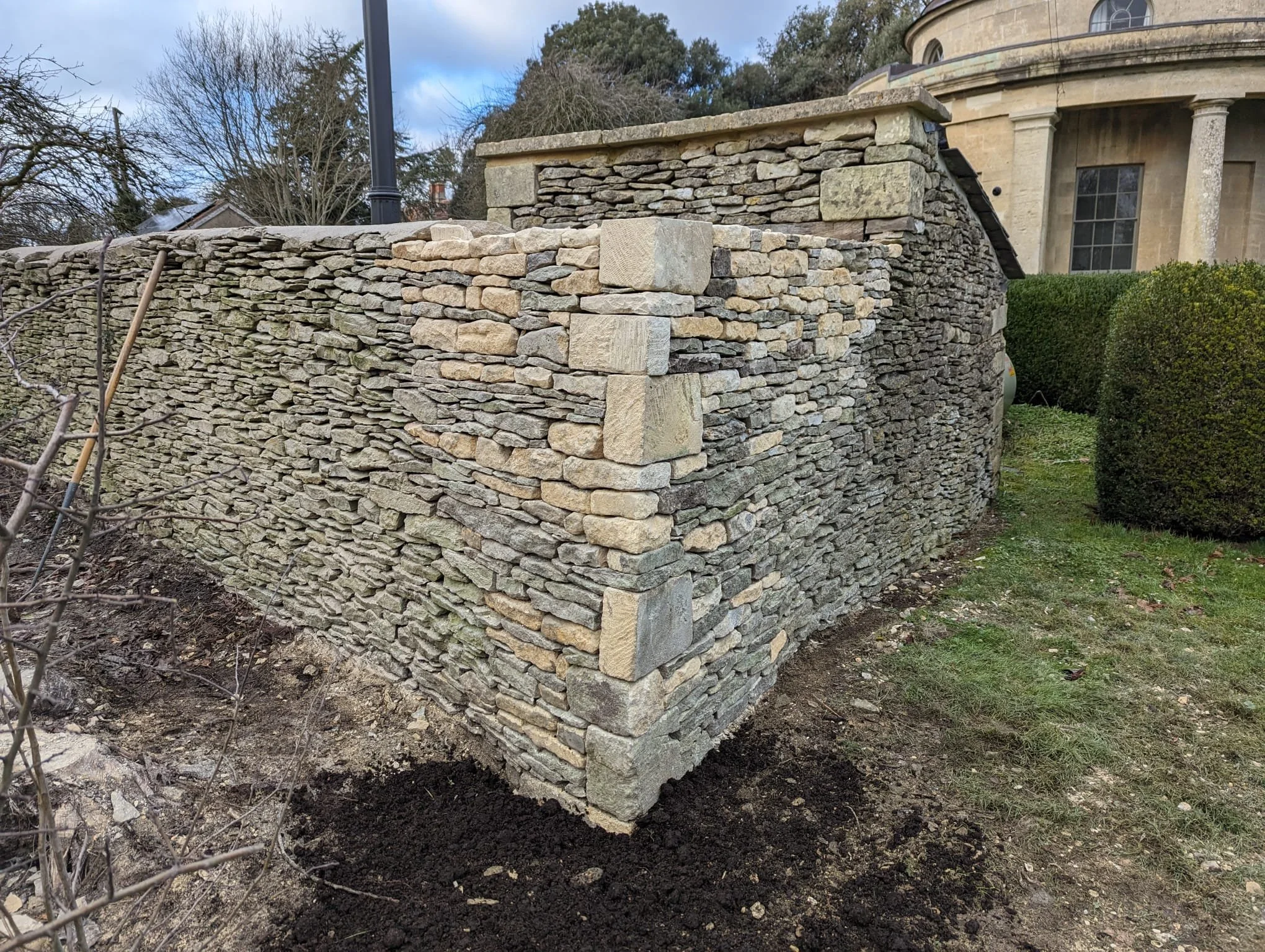 A stone retaining wall constructed with flat, irregularly shaped stones featuring a corner with larger, rectangular stones. The wall is situated outdoors near a house with a rounded exterior and circular windows. There are bushes and some bare trees around, and a patch of dark soil at the base of the wall.
