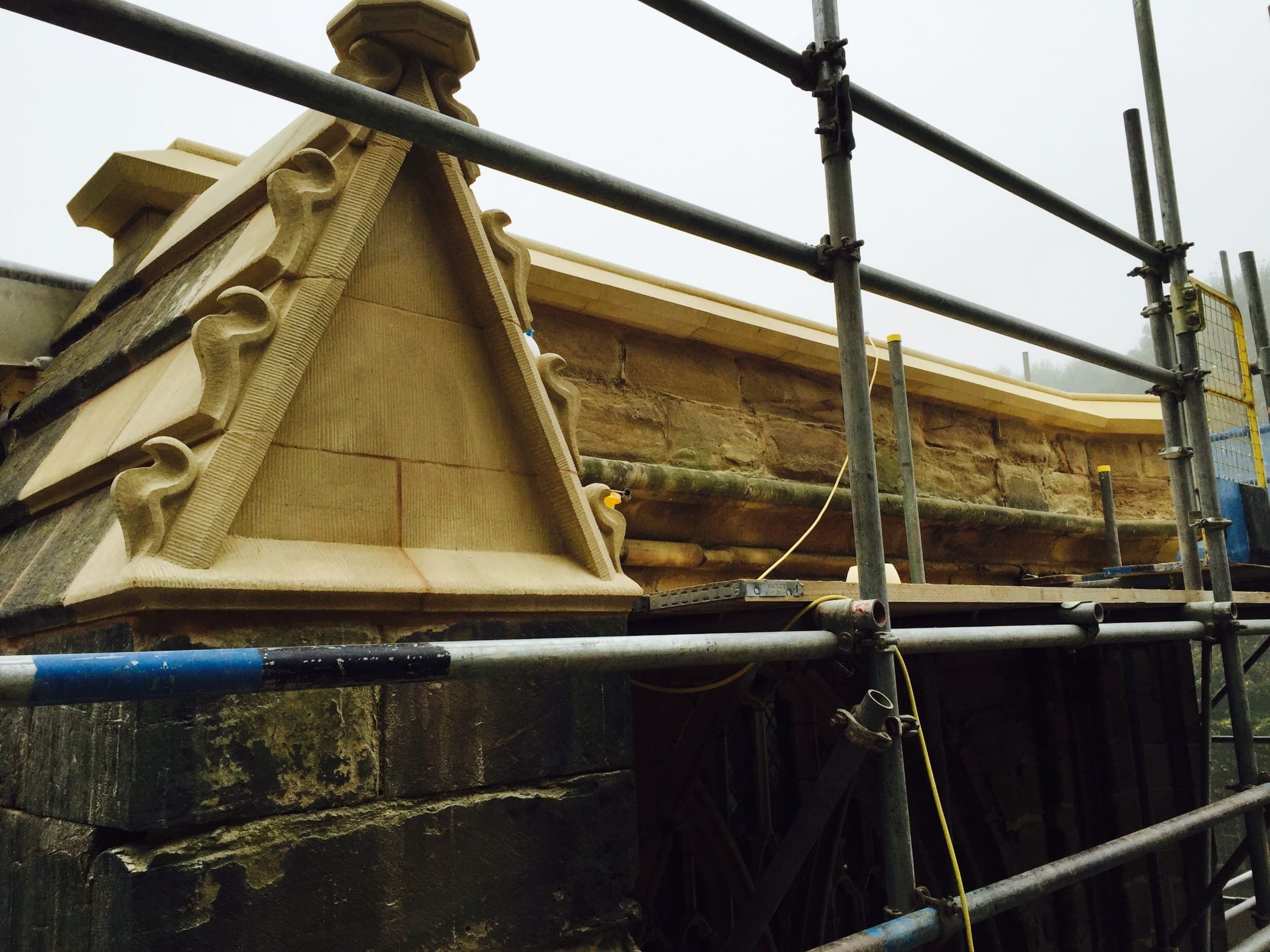 Construction scaffolding surrounds a stone building with decorative stonework on the roof edge. The roof appears to be under renovation or repair.