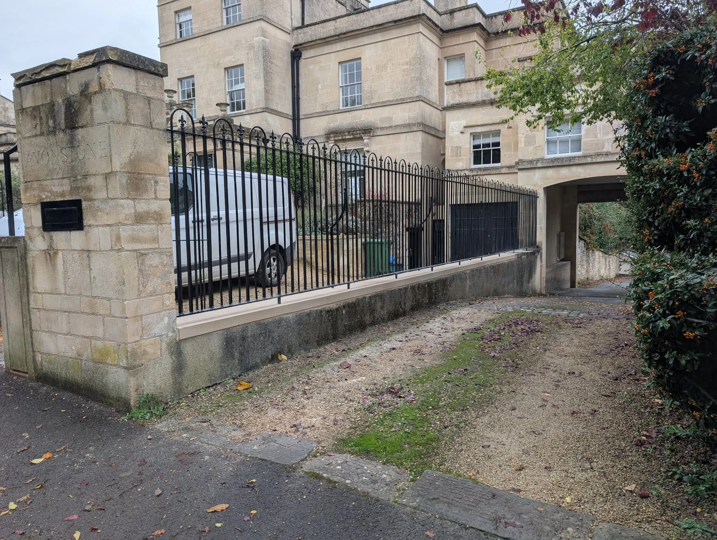 A stone and metal fence surrounds a parking area with a white van parked inside. Behind the fence, a multi-story stone building with several windows is visible. The ground outside the fence is a mix of dirt and moss, with a small alleyway leading under the building.