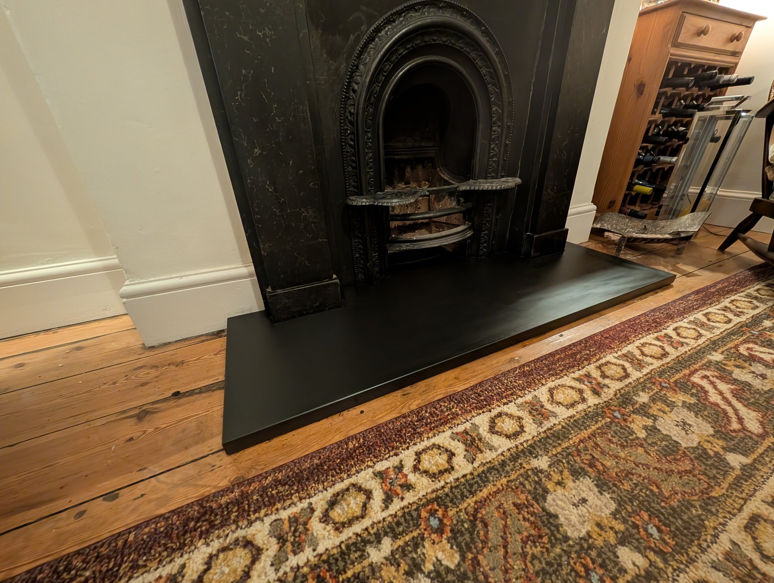 Black fireplace with ornate interior, sitting on a black hearth in front of a white wall, next to a patterned rug on wooden floor, with a wine rack and other furniture nearby.