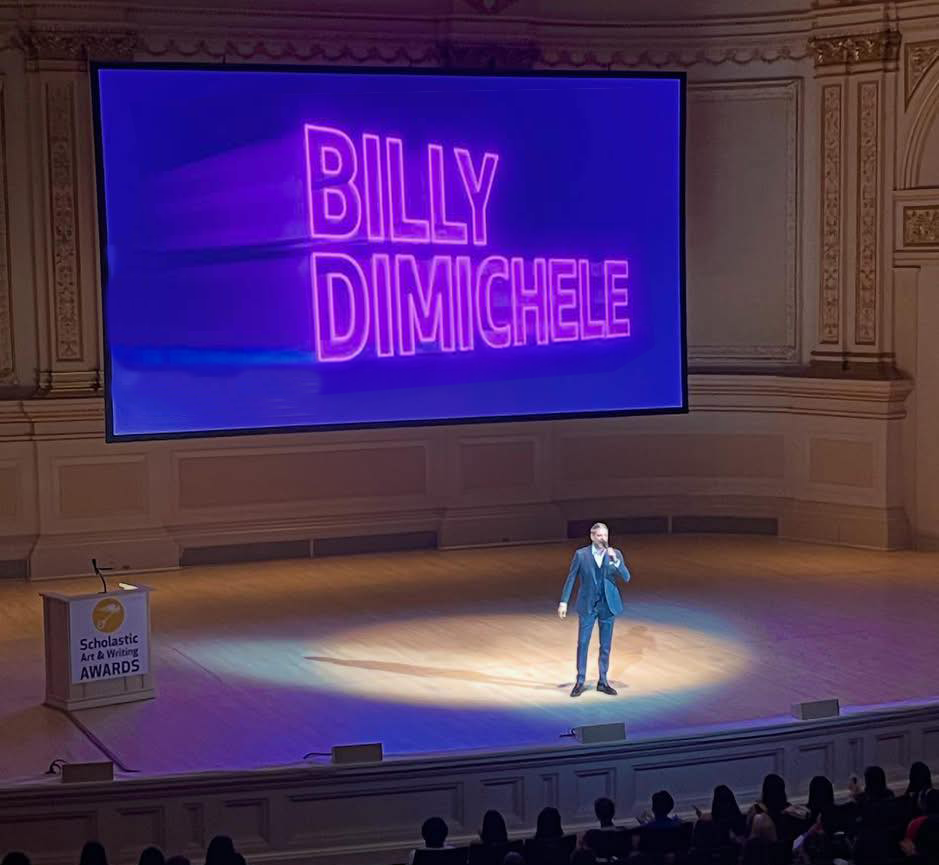 Billy DiMichele is performing at Carnegie Hall at the Scholastic Art & Writing Awards, with his name displayed in large purple neon letters on a screen behind him. The stage is lit with a spotlight, and an audience is seated in front.