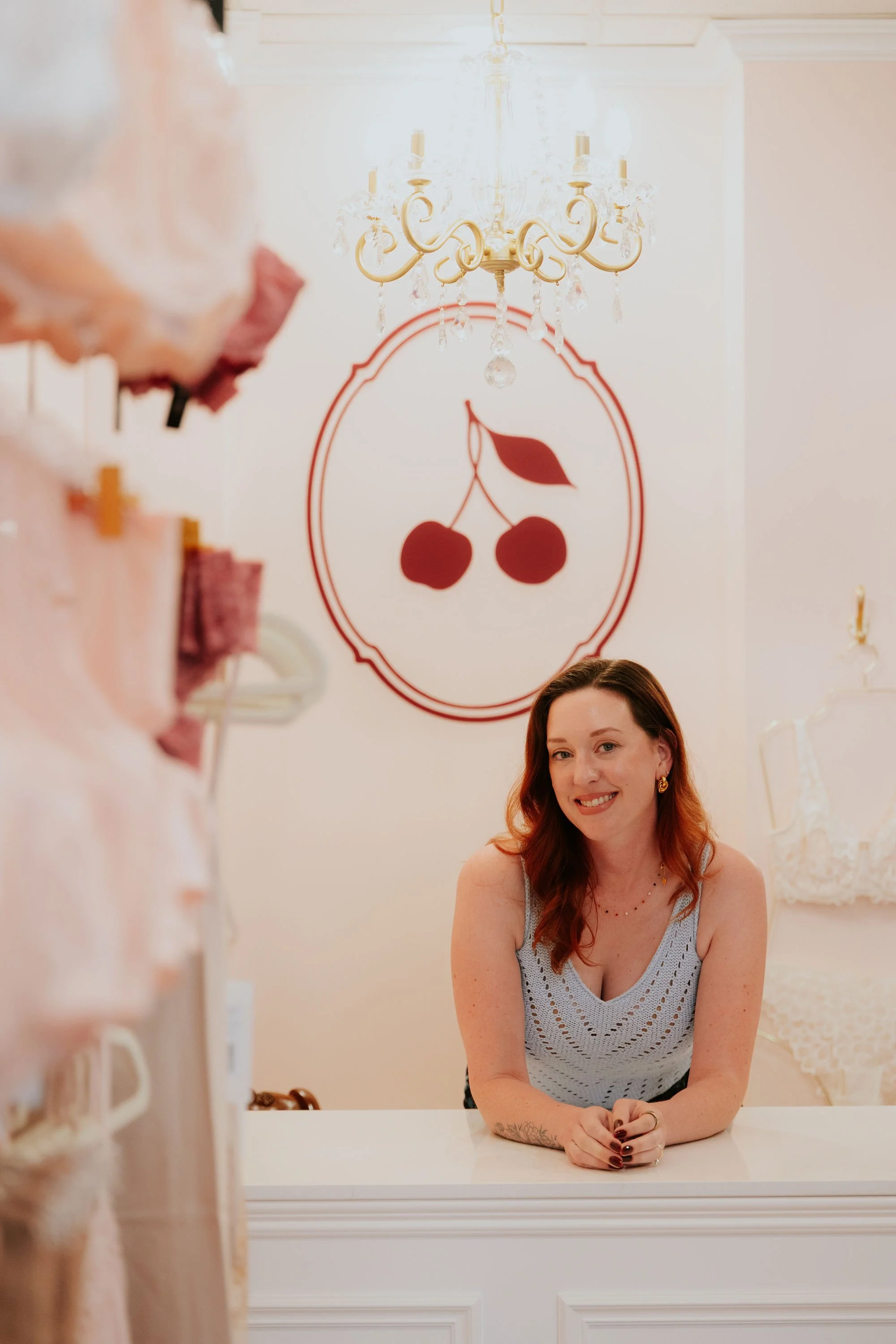A woman with red hair is sitting at a white counter with her hands clasped, smiling, in a room decorated with a chandelier and a framed cherry logo on the wall.