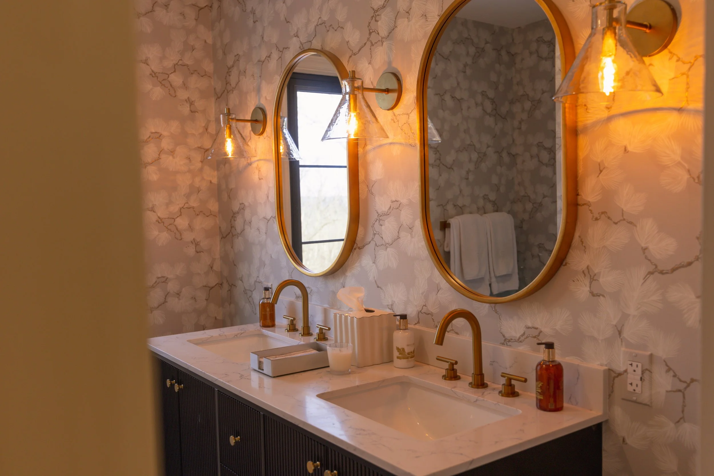 Bathroom with two oval mirrors, wall-mounted lights with glass shades, marble countertop with dual sinks, soap dispensers, tissue box, and a window with a view outside. Towels are hanging on a rail reflected in the right mirror.
