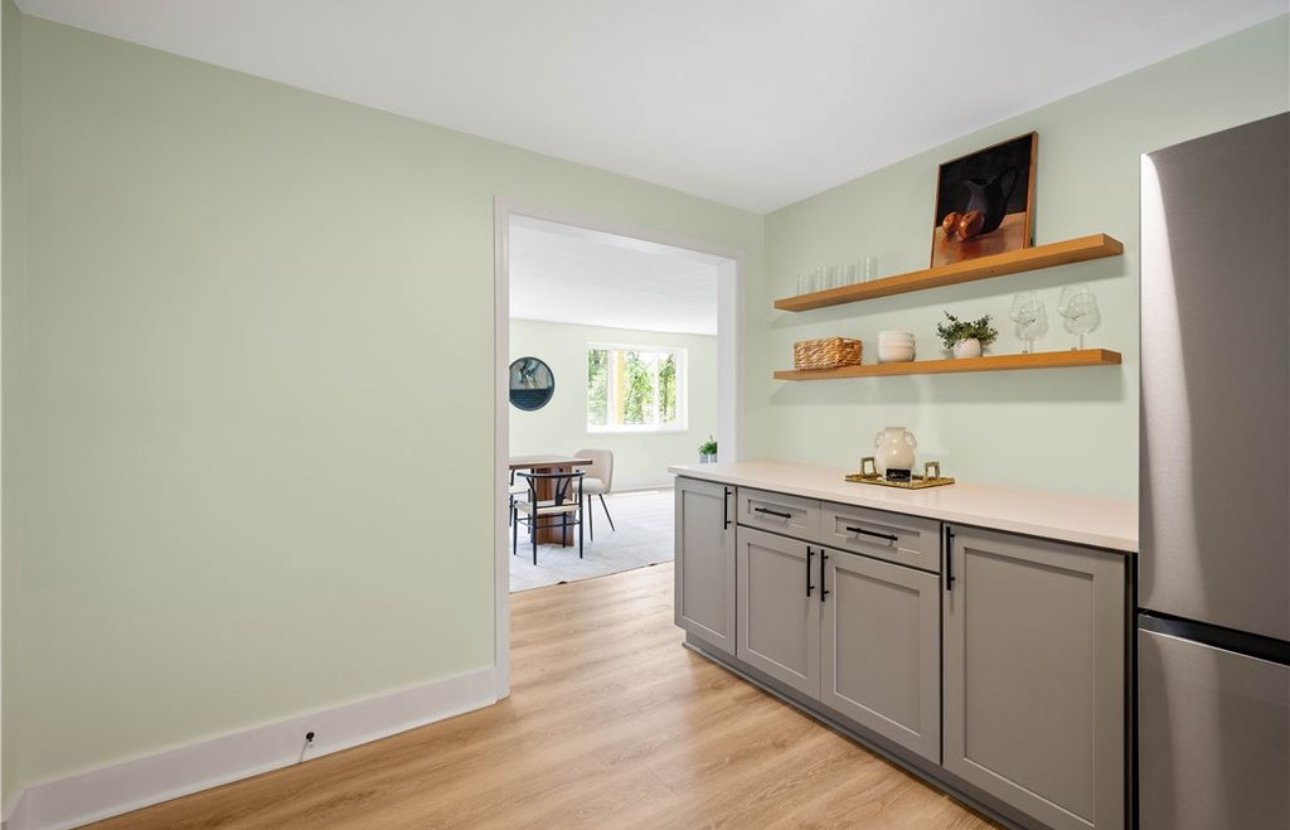 Kitchen with light green walls, gray cabinets, wooden open shelves, and a stainless steel refrigerator, with a view into a dining area with windows and seating.