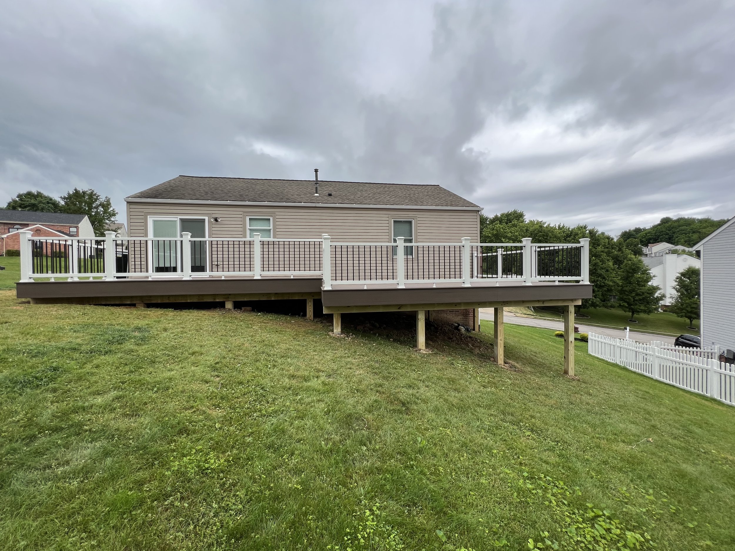 Backyard view of a house with a newly built deck with white and black railing, on a slightly sloped grassy yard under a cloudy sky.