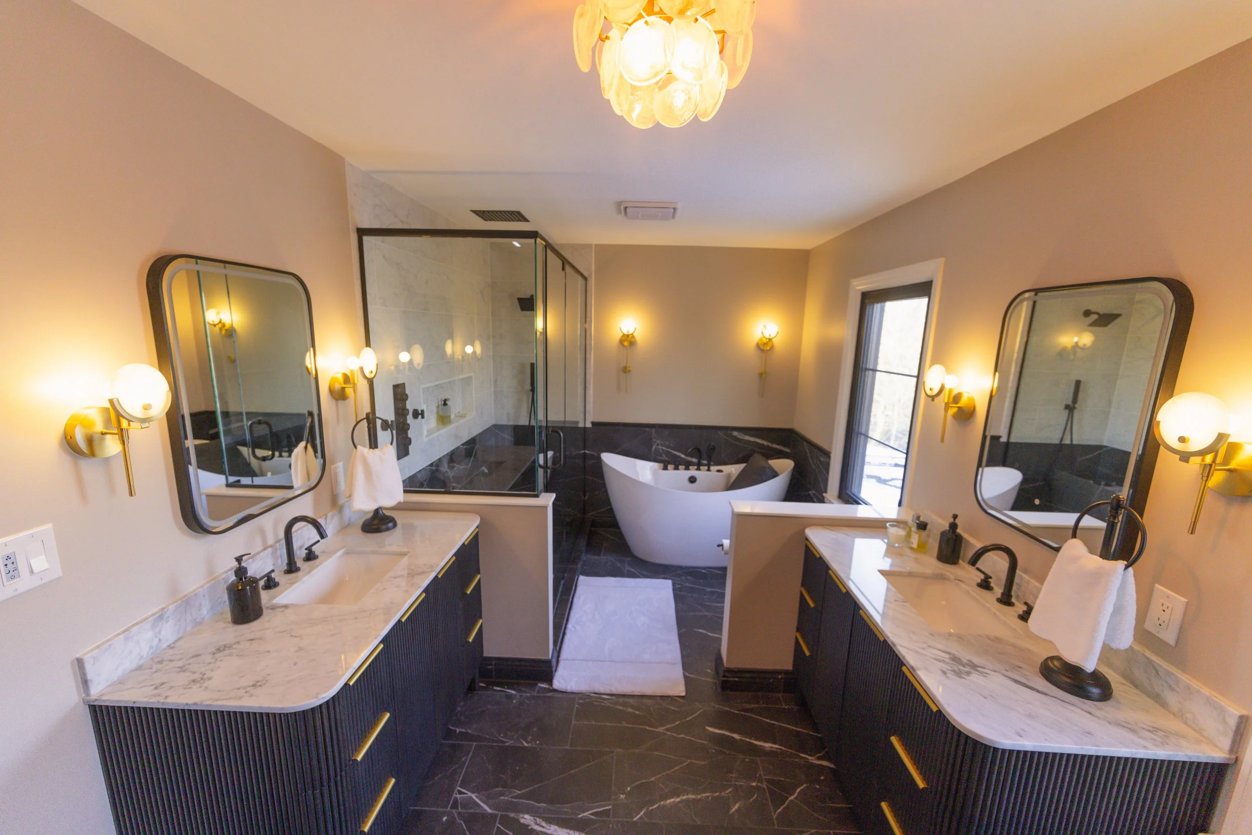 Modern bathroom featuring dual sinks with marble countertops, large mirrors, wall-mounted lights, a glass-enclosed shower, a free-standing bathtub, and a large window with natural light.