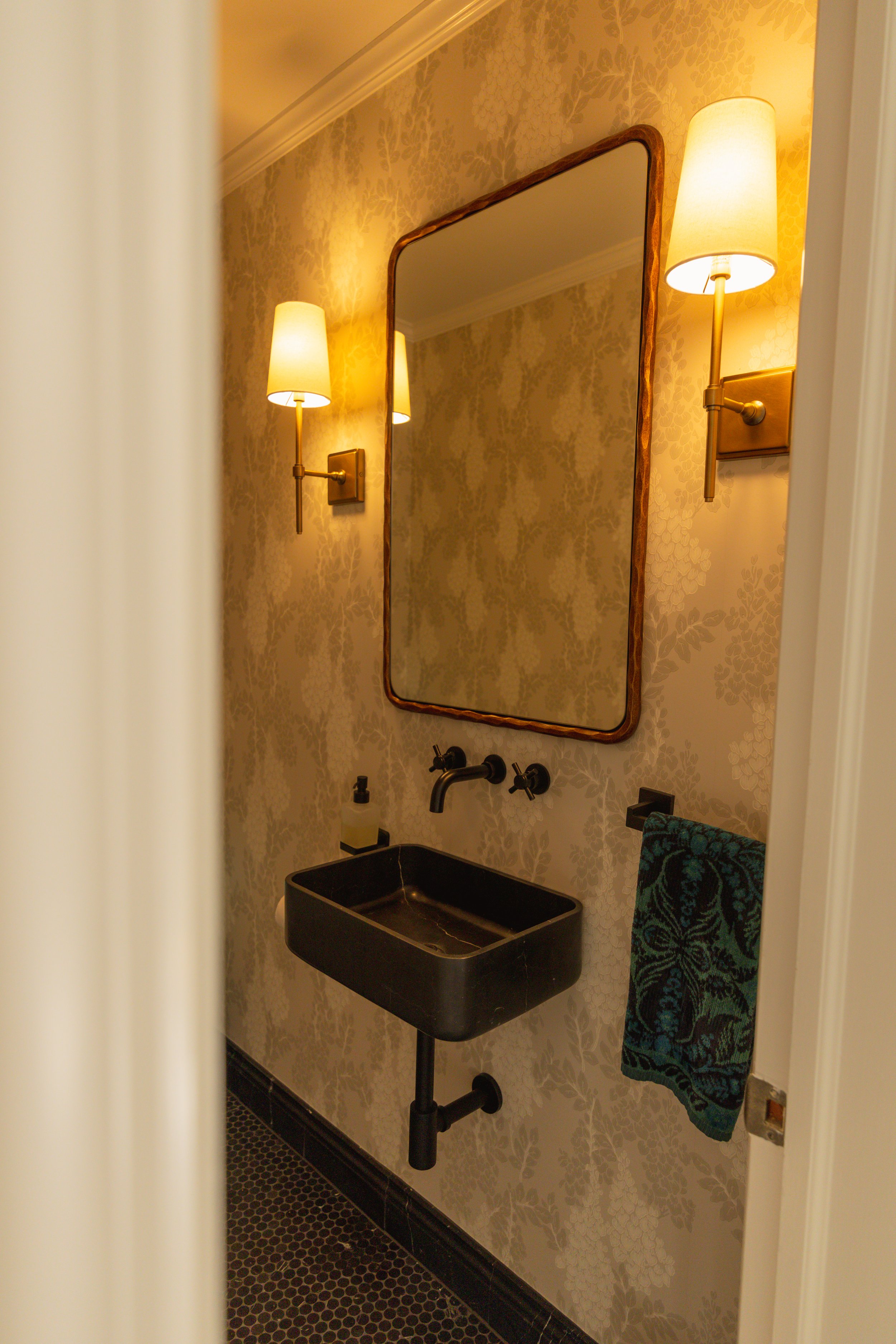A small bathroom with a black wall-mounted sink, a mirror, and two wall sconces with cream-colored lampshades on either side of the mirror. The wall has a patterned wallpaper and there is a teal towel hanging on a hook to the right of the sink.