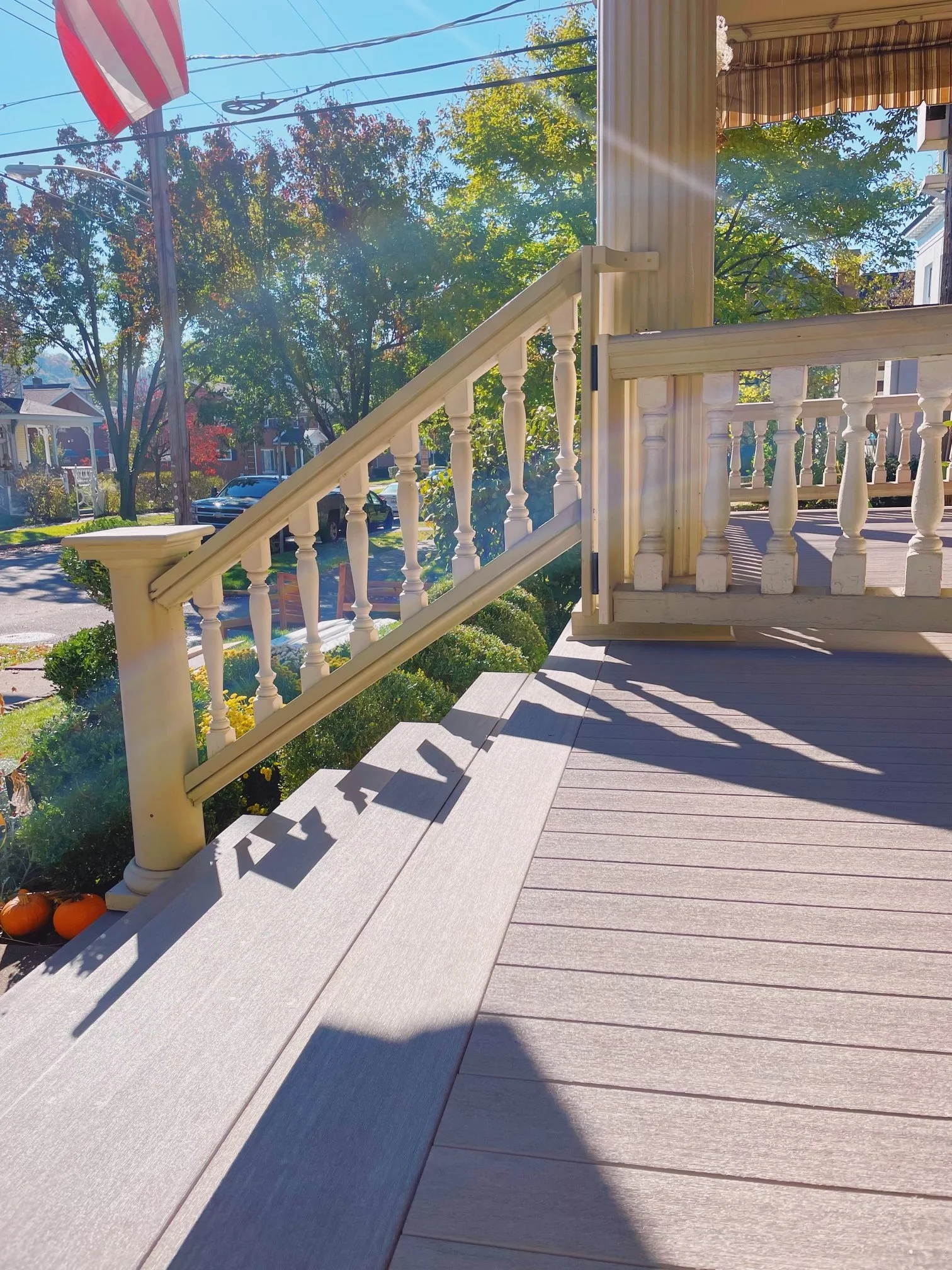 A porch with wooden flooring, white railing, and stairs leading down. Shadows cast by the railing and trees, with a suburban street and trees in the background.
