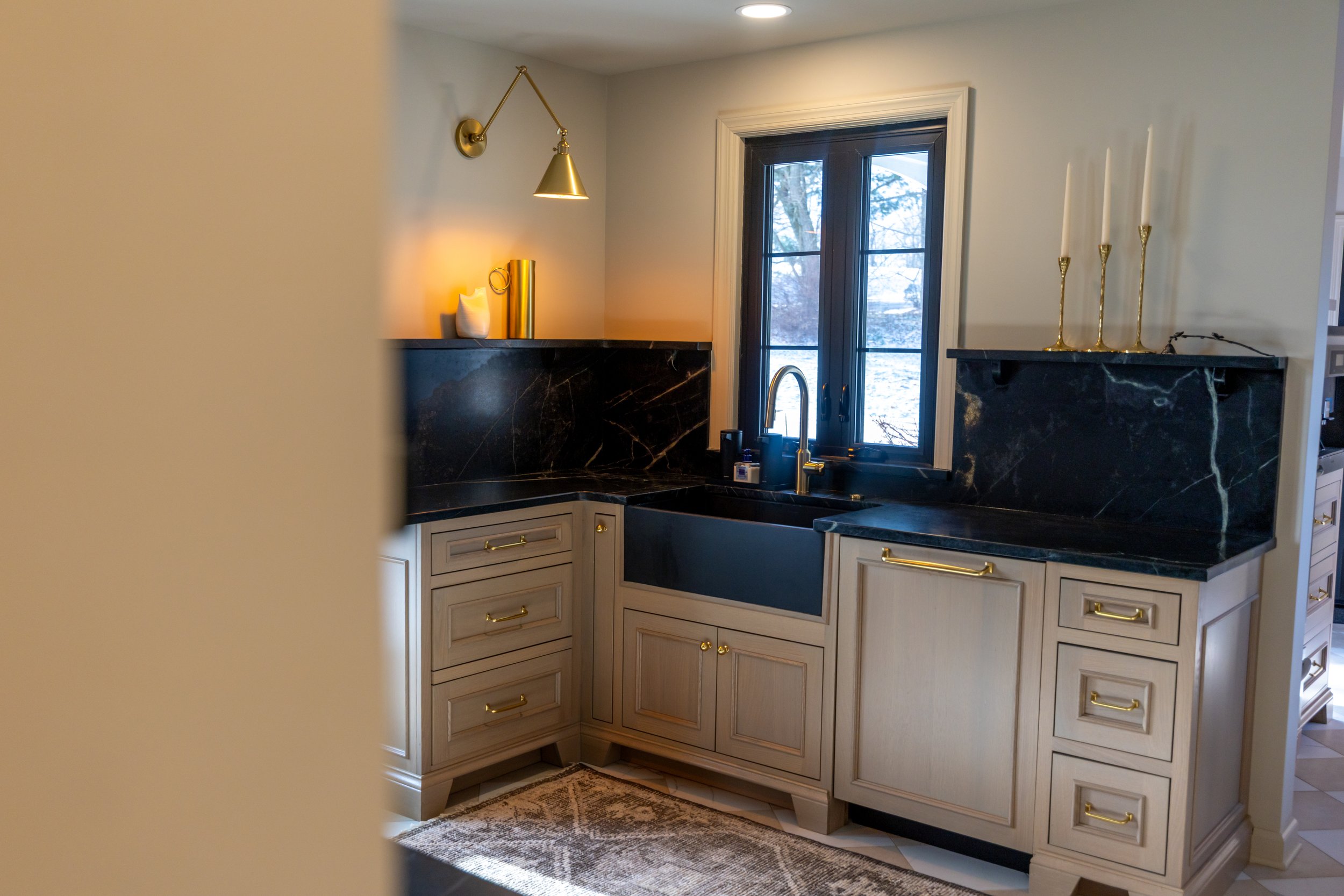Kitchen with beige cabinetry, black marble countertops, and a black marble backsplash, featuring a black farmhouse sink, gold hardware, and a window overlooking snowy outdoors with decorative gold candlesticks and a gold sconce on the wall.