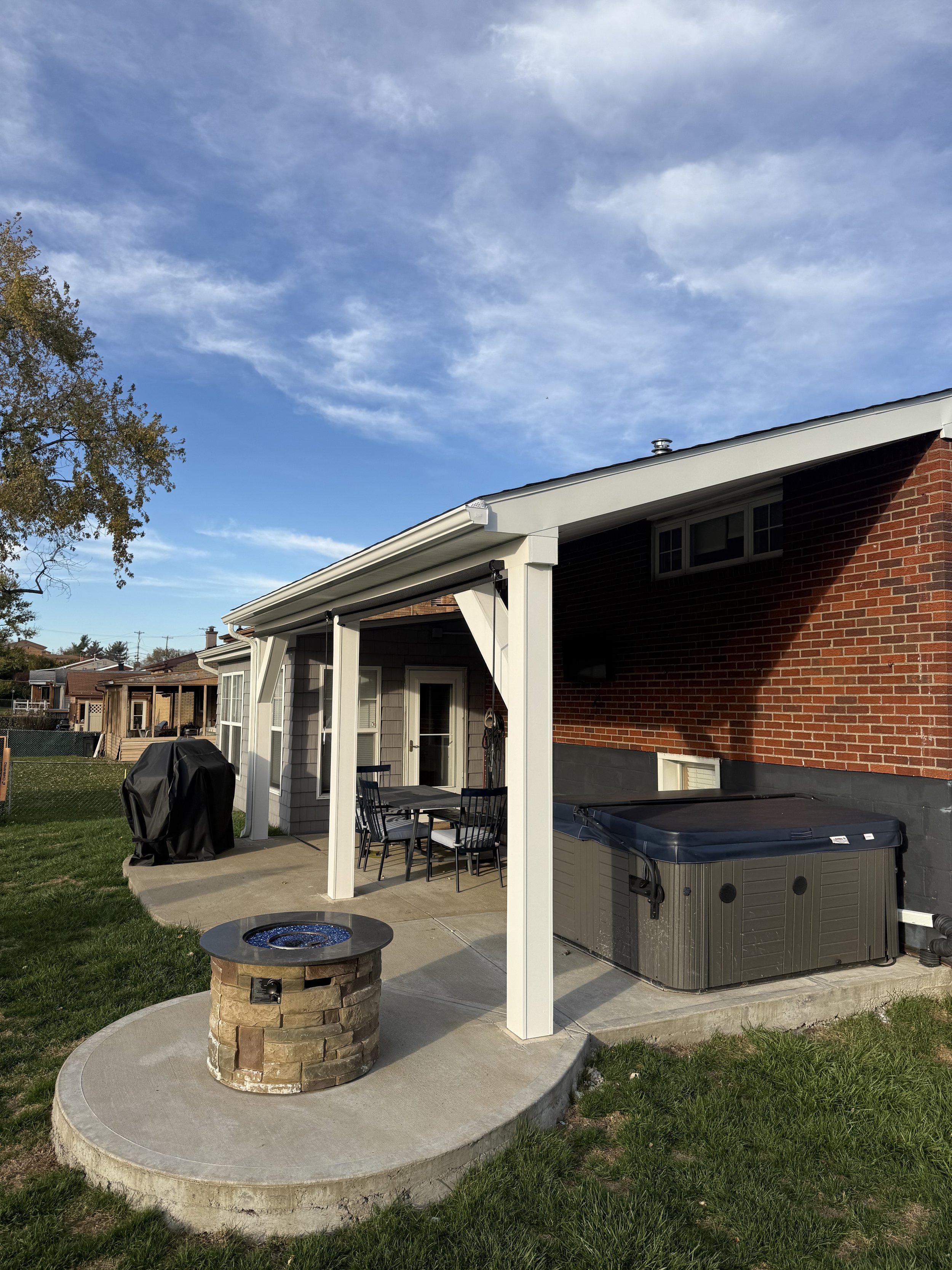 Backyard patio with a concrete slab, a fire pit, an outdoor dining table with chairs, a grill, a hot tub, and a house with a brick and siding exterior, under a blue sky with some clouds.