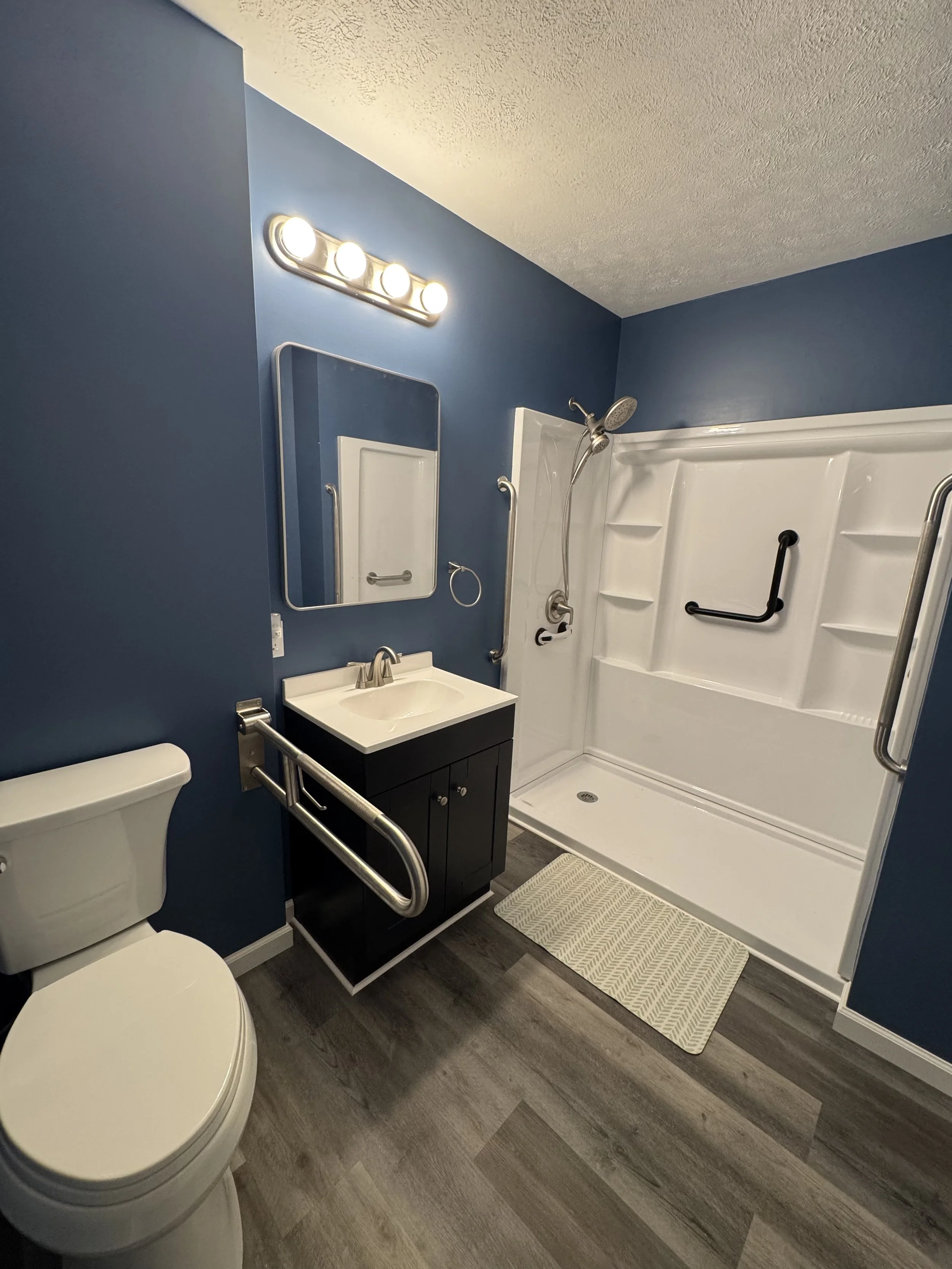 A bathroom with blue walls, a white toilet, a black vanity with a white sink, a mirror, a wall-mounted light fixture, and a walk-in shower with grab bars and a textured beige mat.