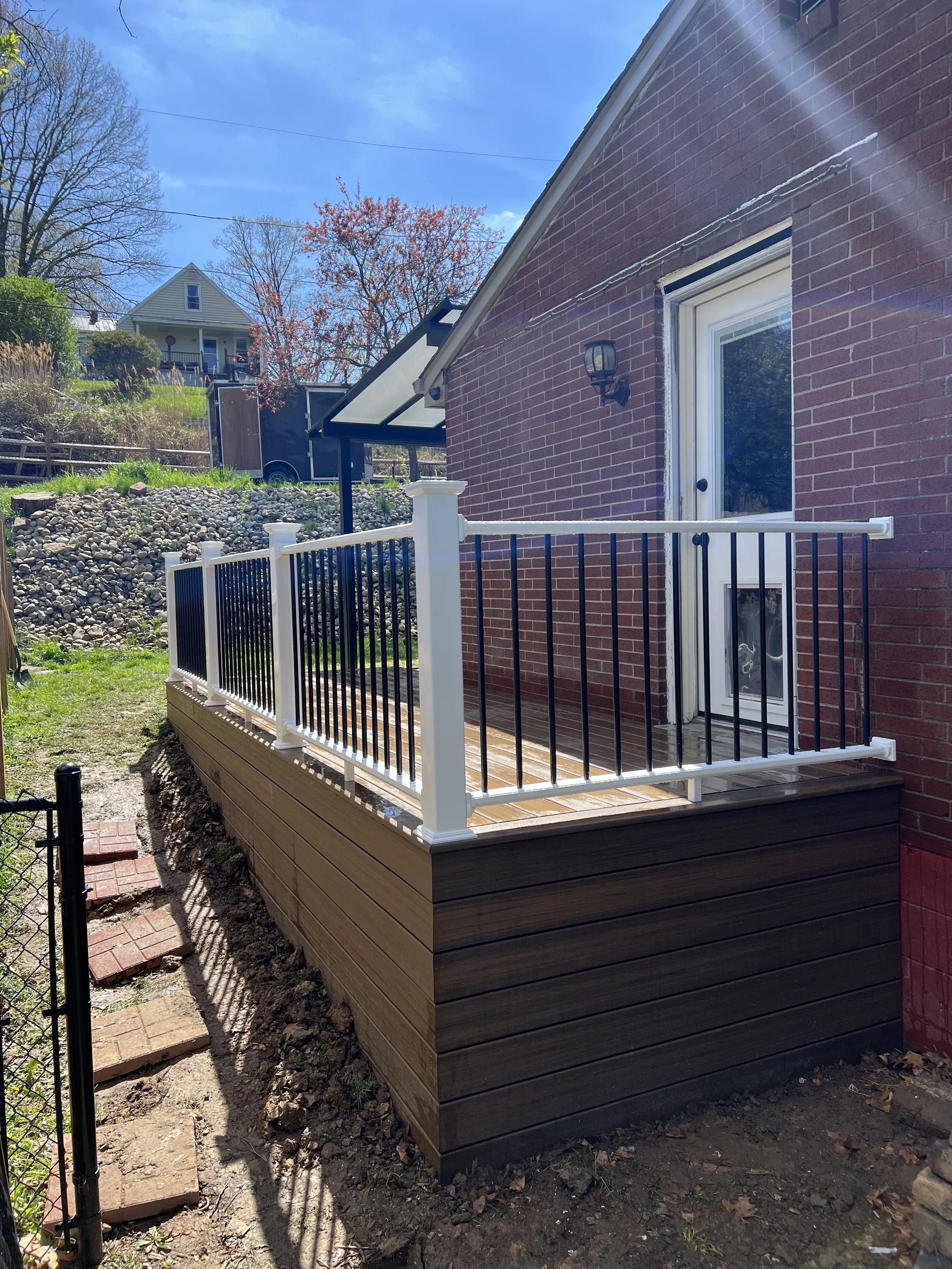 Newly built small outdoor deck with brown wooden flooring and white and black metal railing, attached to a brick house with a sliding glass door, outdoor light fixture, and a small black hook on the wall.